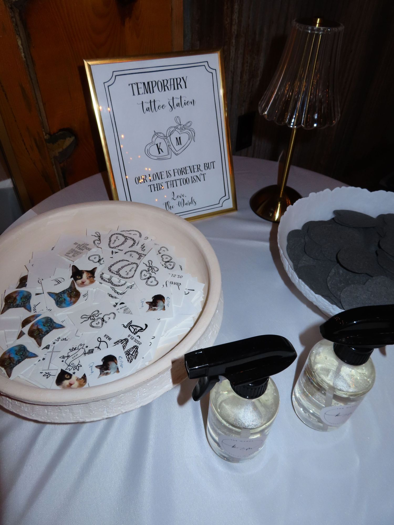 Temporary tattoo station setup at a wedding with decorative tray, spray bottles, and framed sign on white tablecloth.
