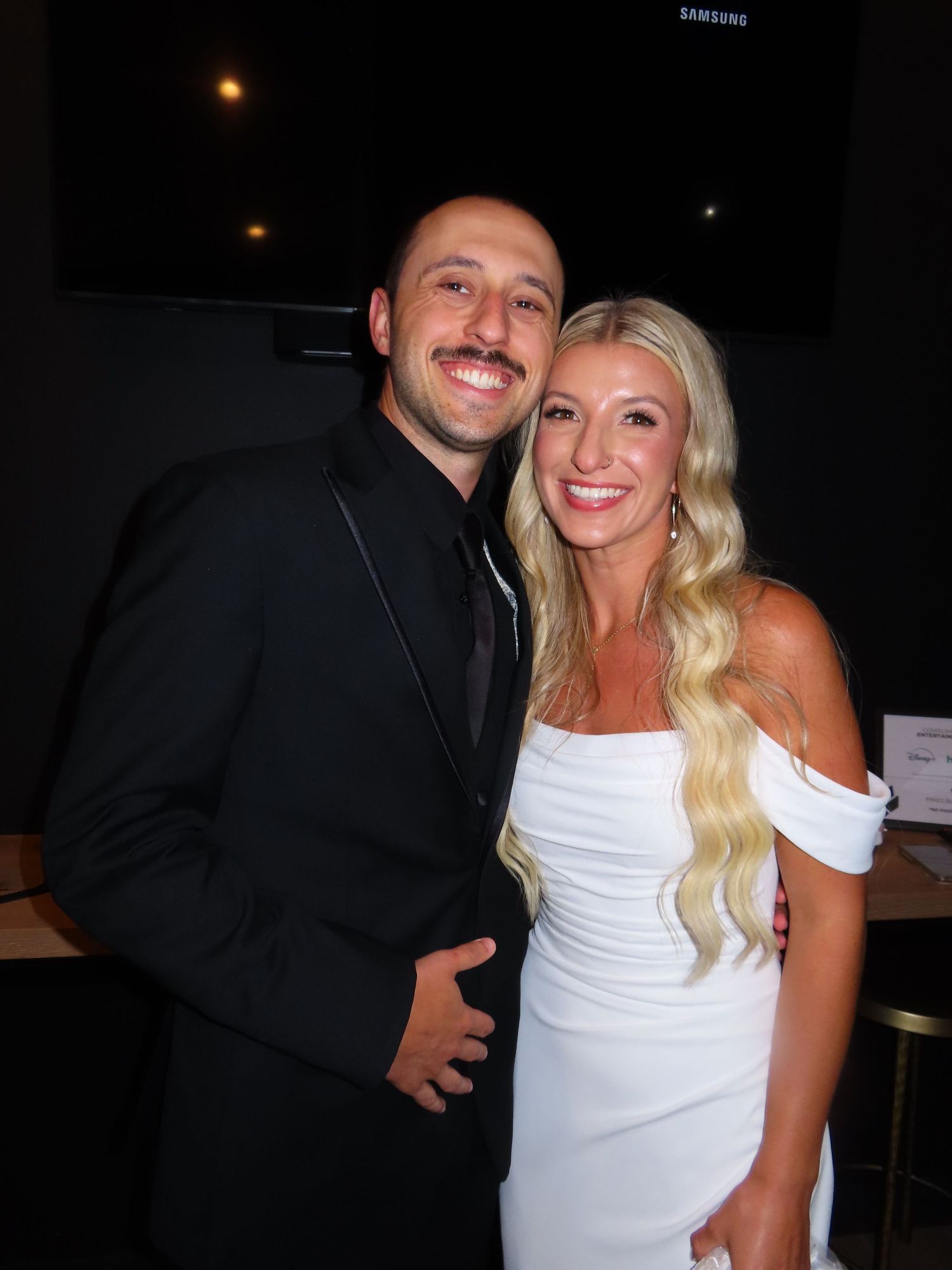 Smiling couple posing together at event, man in black suit and woman in white dress.