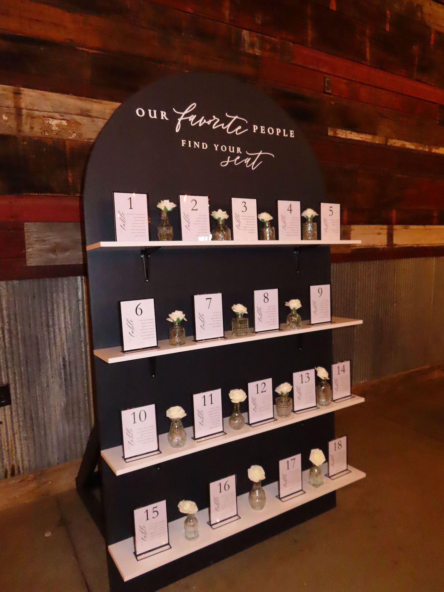 Elegant wedding seating chart display with numbered cards and white roses on a dark arched board reading Our Favorite People Find Your Seat.