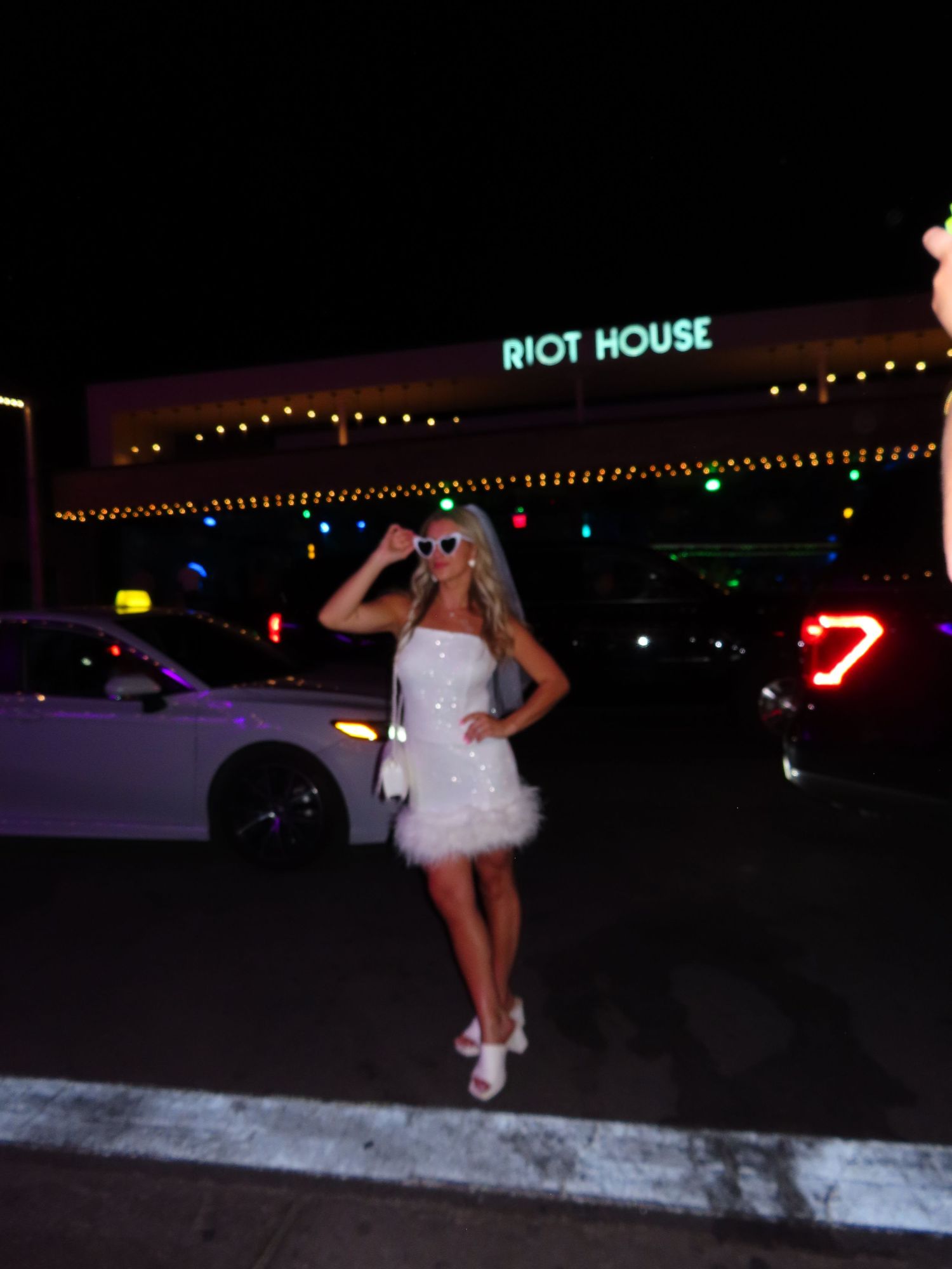 Woman in white feather mini dress and sunglasses posing outside Riot House at night.