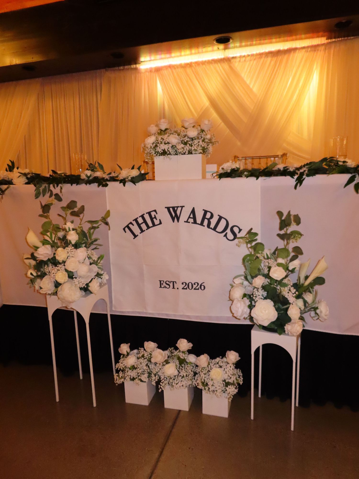 Elegant wedding backdrop for The Wards EST 2026 adorned with white roses, greenery, and floral arrangements on white stands.