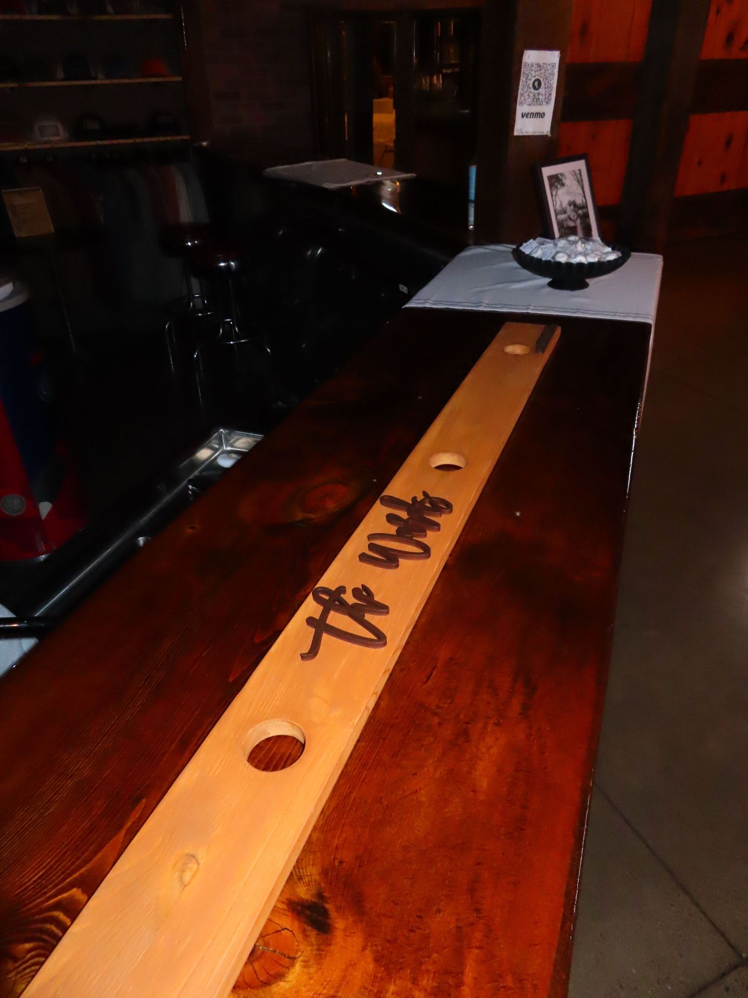 A rustic wooden bar counter with a light wood strip inlay, circular holes, and cursive writing in a dimly lit venue.