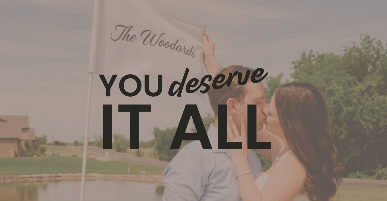 Couple sharing a romantic kiss outdoors at The Woodards venue with text overlay reading You Deserve It All.
