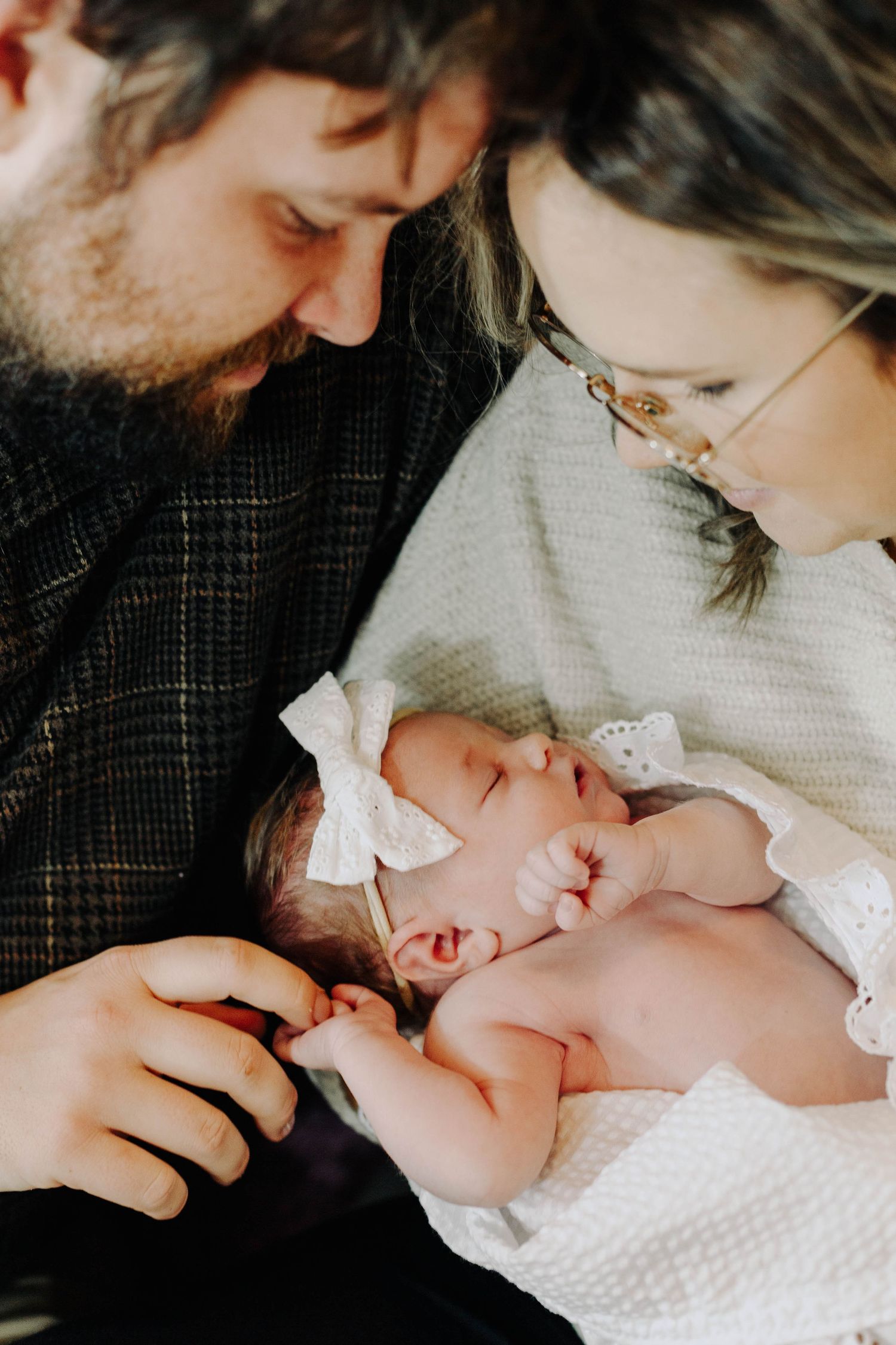 Bearded father and glasses-wearing mother lovingly gaze at their sleeping newborn baby girl with a bow headband.