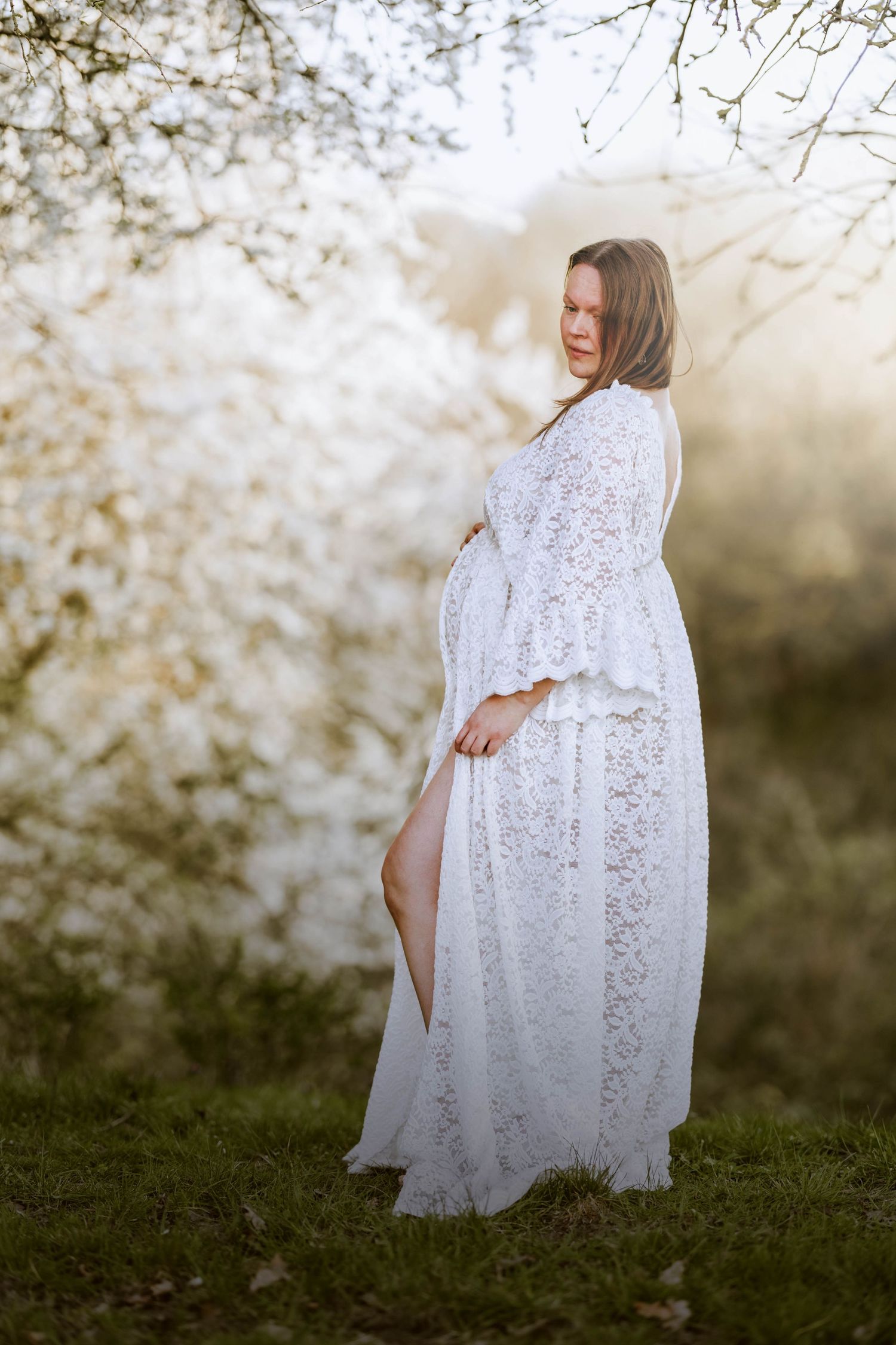 Elegant maternity photo featuring woman in white lace dress amid spring blooms.