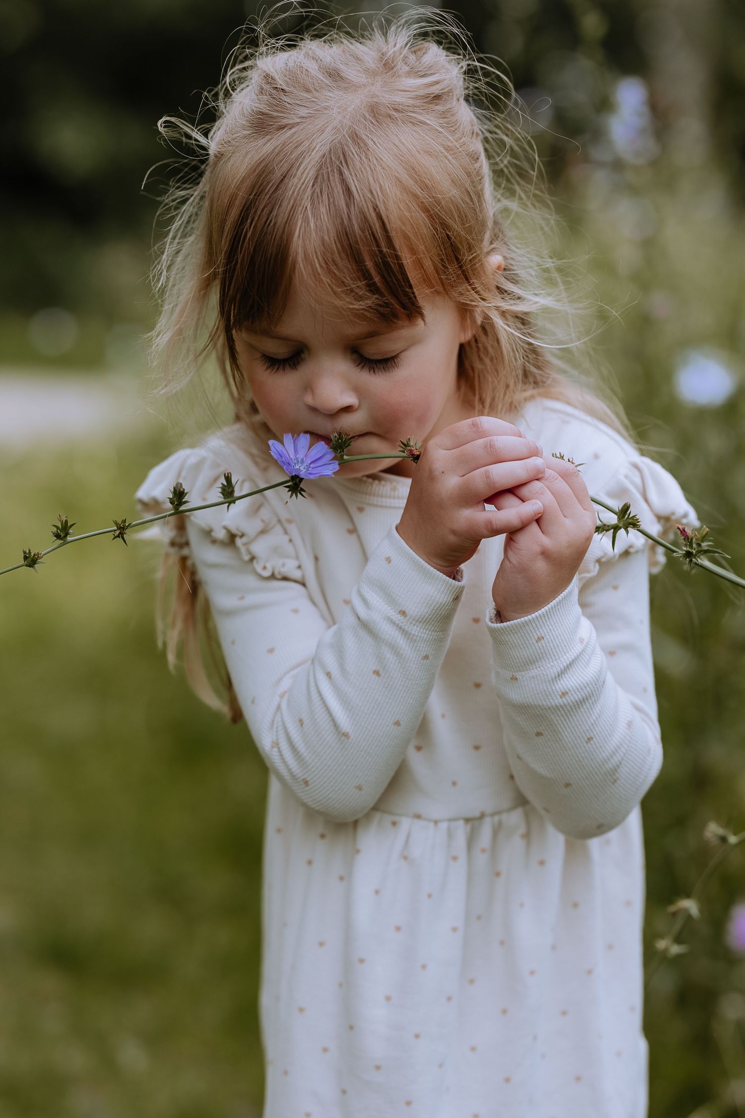 A young child in a white polka dot dress plays shyly in a garden setting with wildflowers.