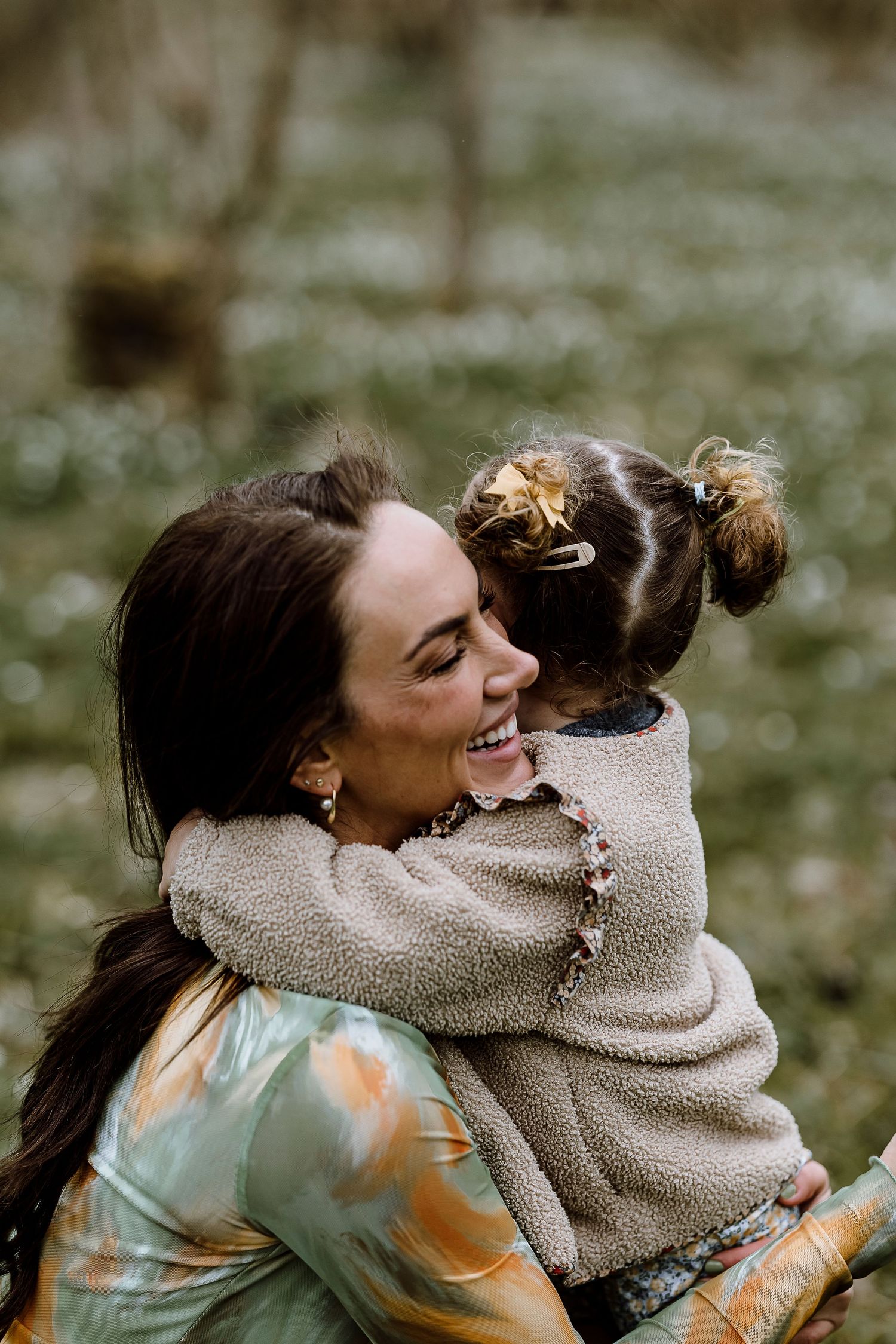 Two people share a joyful embrace outdoors while wearing cozy sweaters on a blurred natural background.