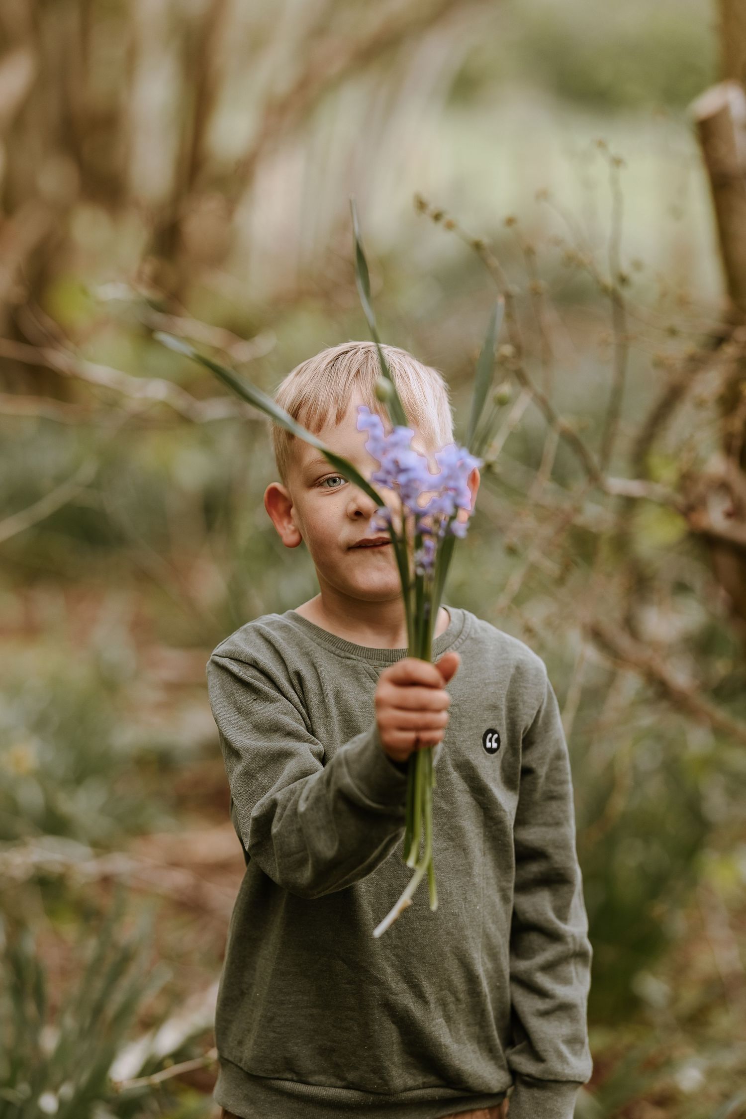 Young child in gray sweater holding up purple wildflowers in an outdoor setting.