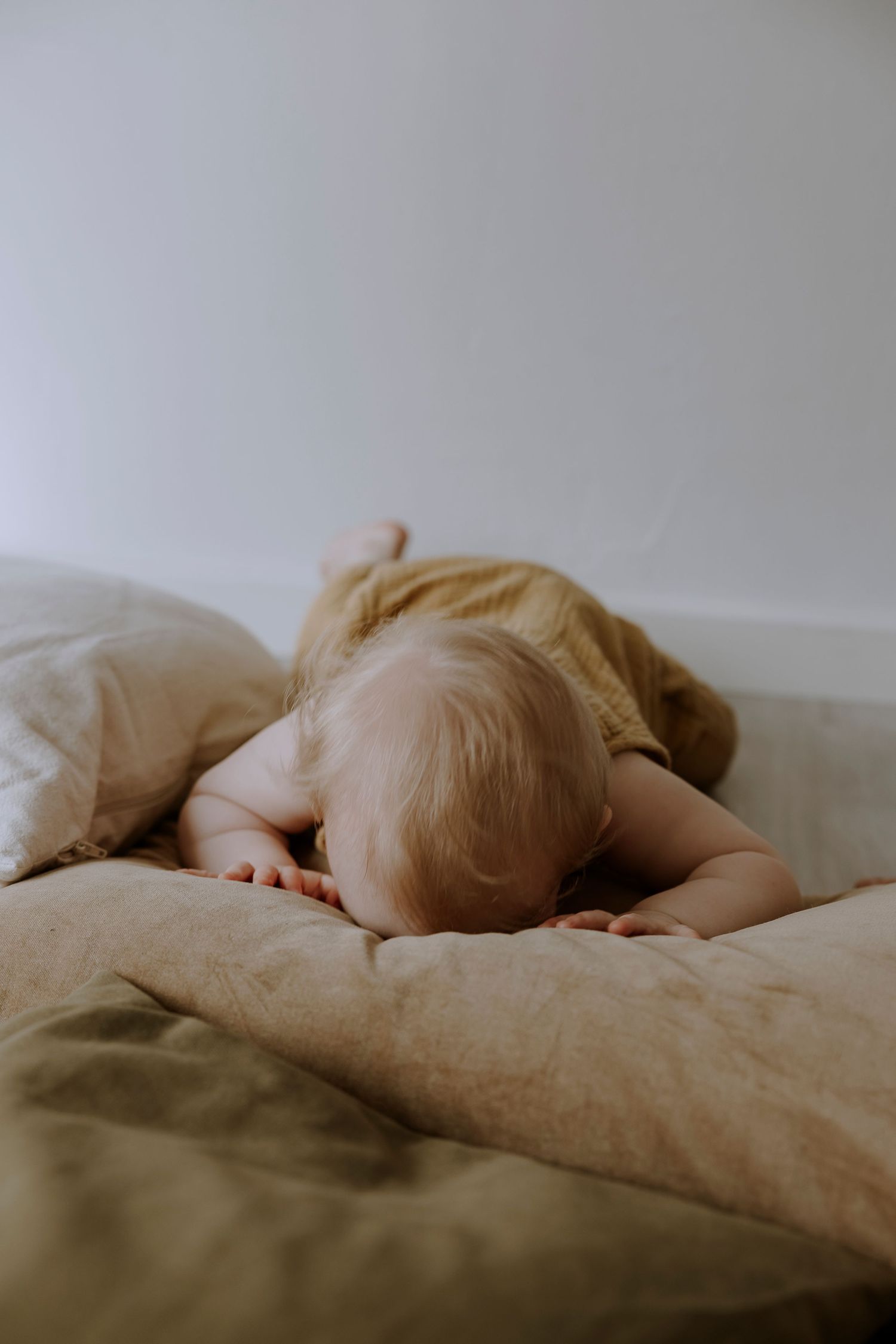 Baby in yellow outfit rests face down on beige pillows.