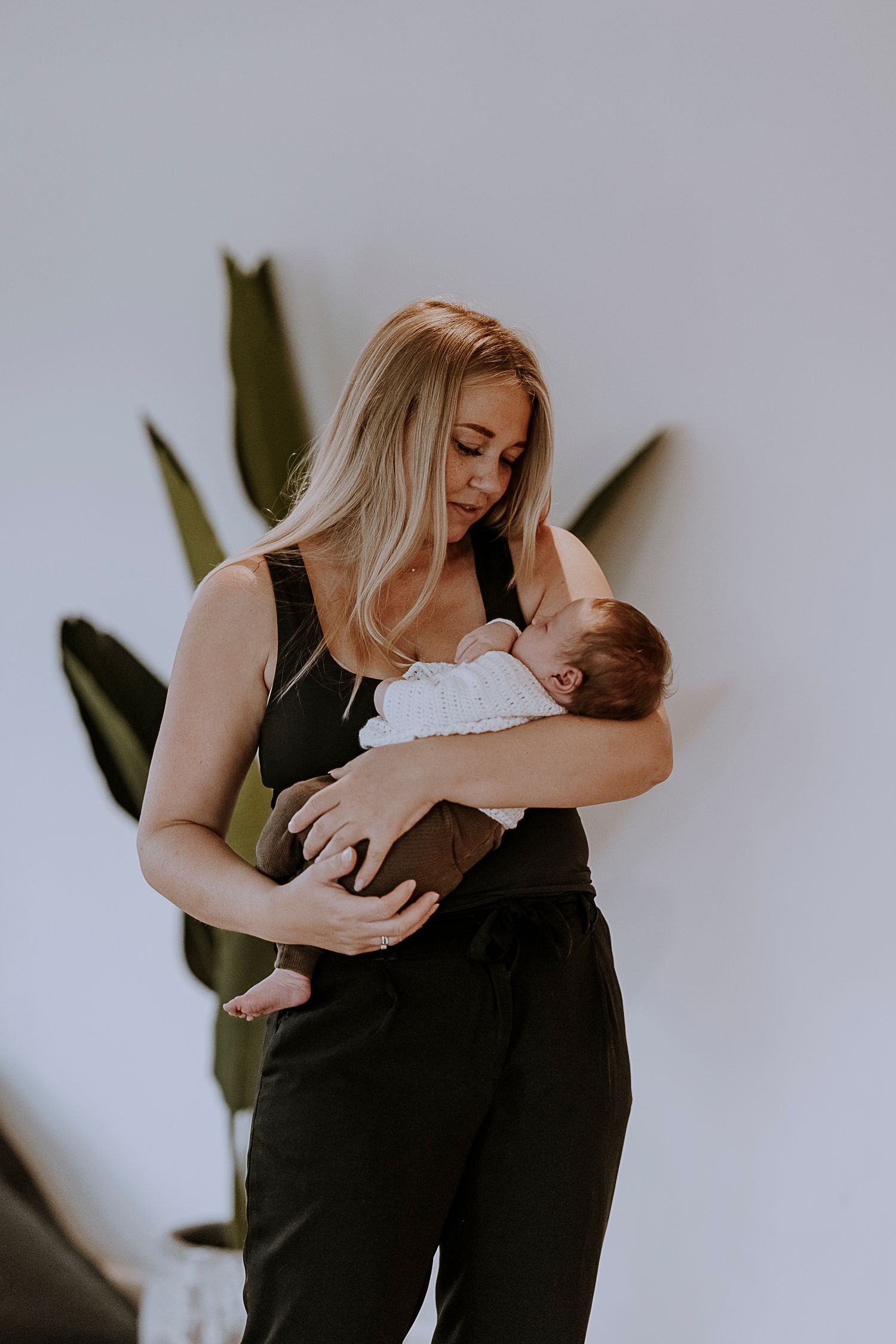 Parent in black outfit holding sleeping newborn against white wall with plant.