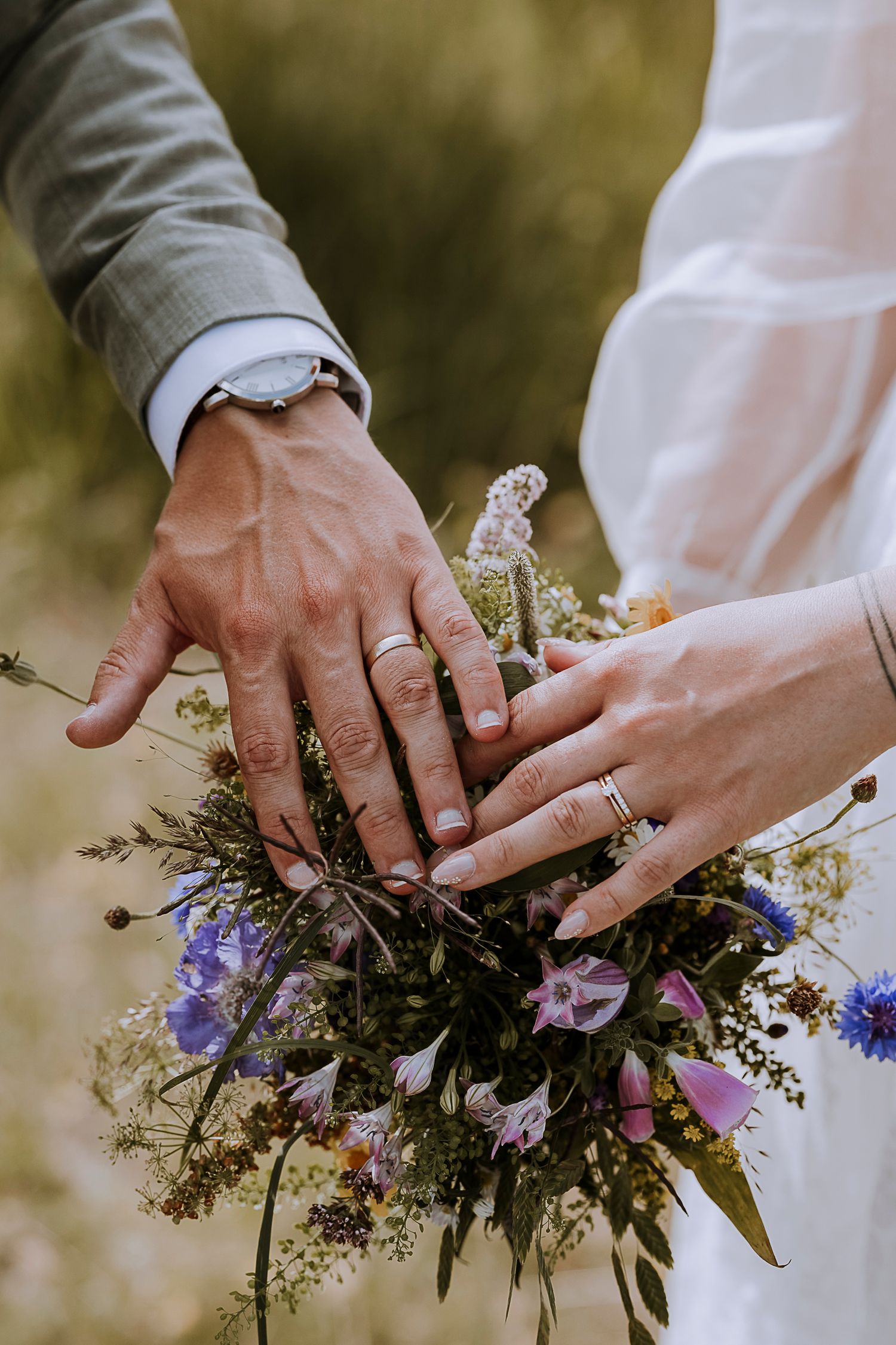 Close-up of newlywed hands with rings resting on wildflower wedding bouquet.