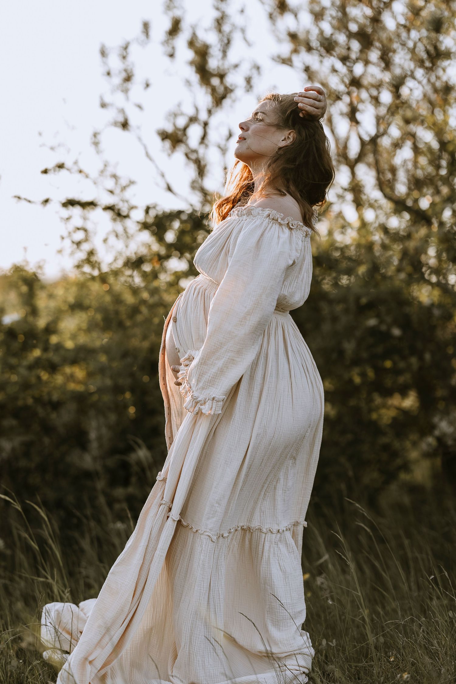 A person in a flowing white vintage dress stands in a sunlit field during golden hour with trees in the background.