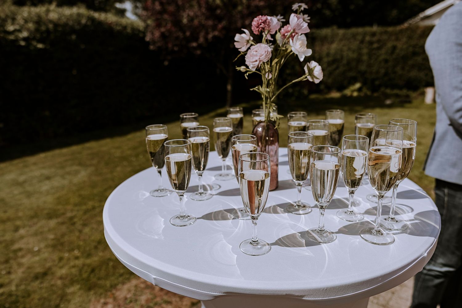 White round table with champagne flutes and pink floral centerpiece at garden reception.