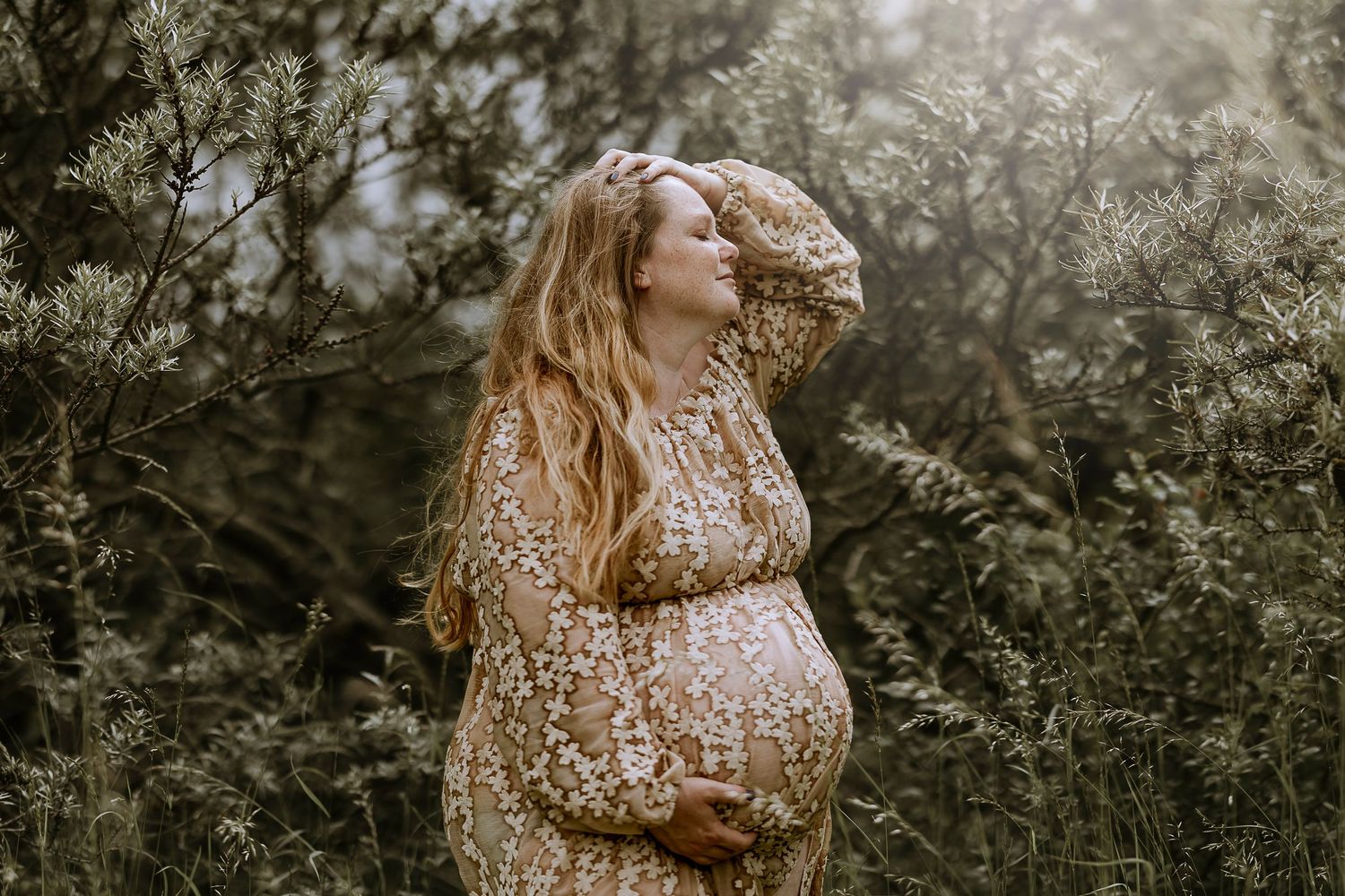 A pregnant woman in a floral dress poses in nature with sunlight streaming through trees.