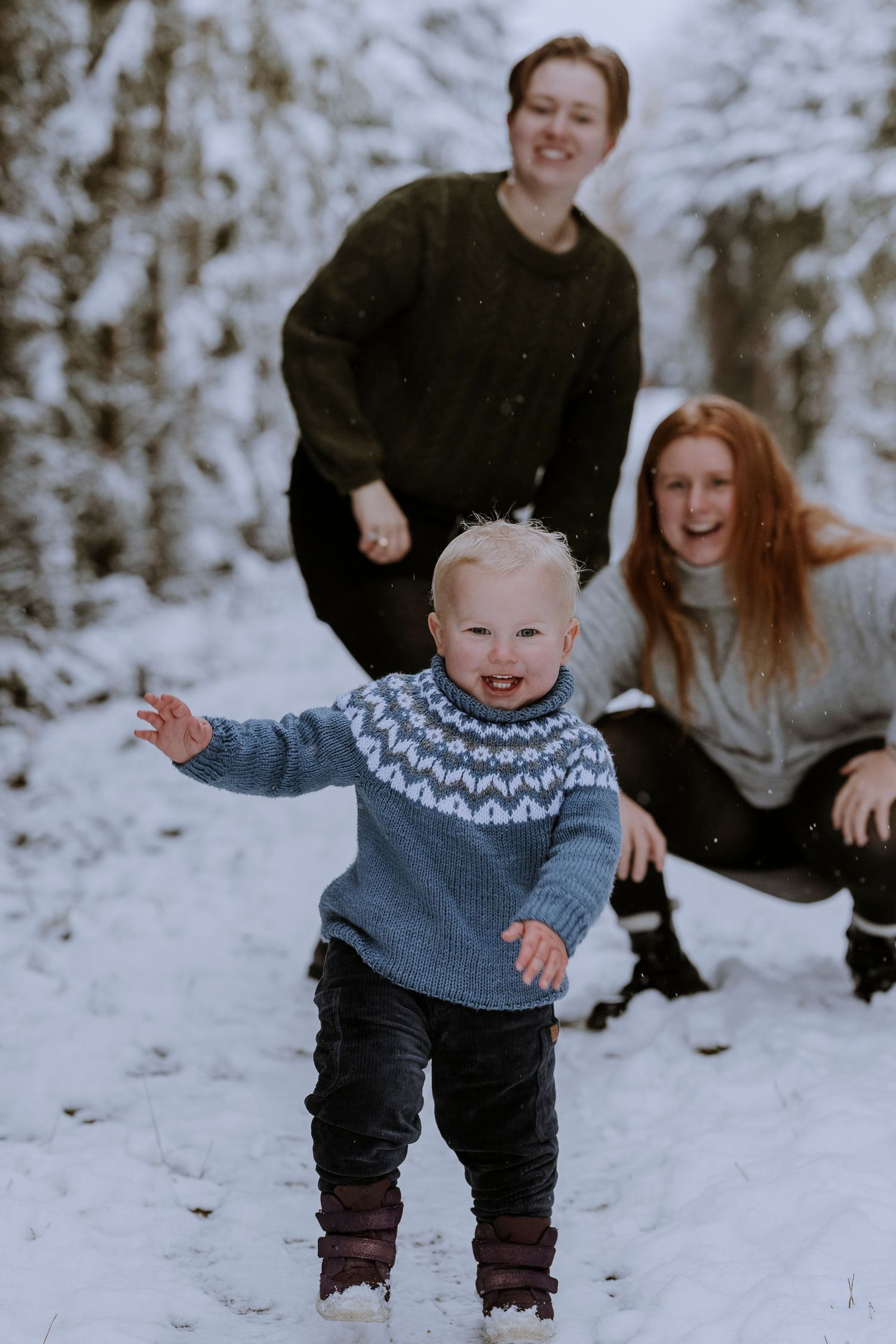 Family enjoying winter snow day together in forest setting with joyful toddler in blue sweater.