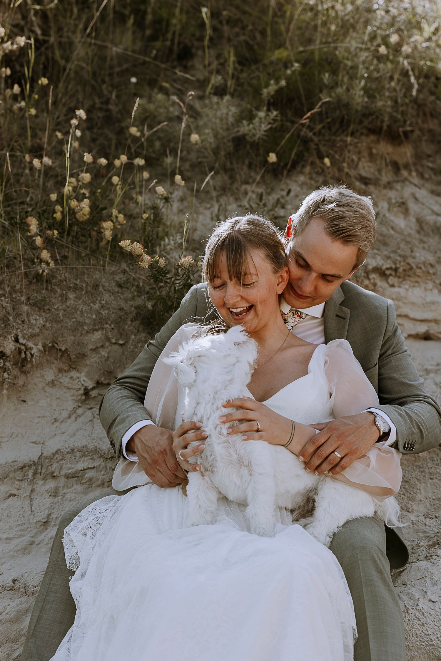Wedding couple laughing together on sandy slope with their white dog.