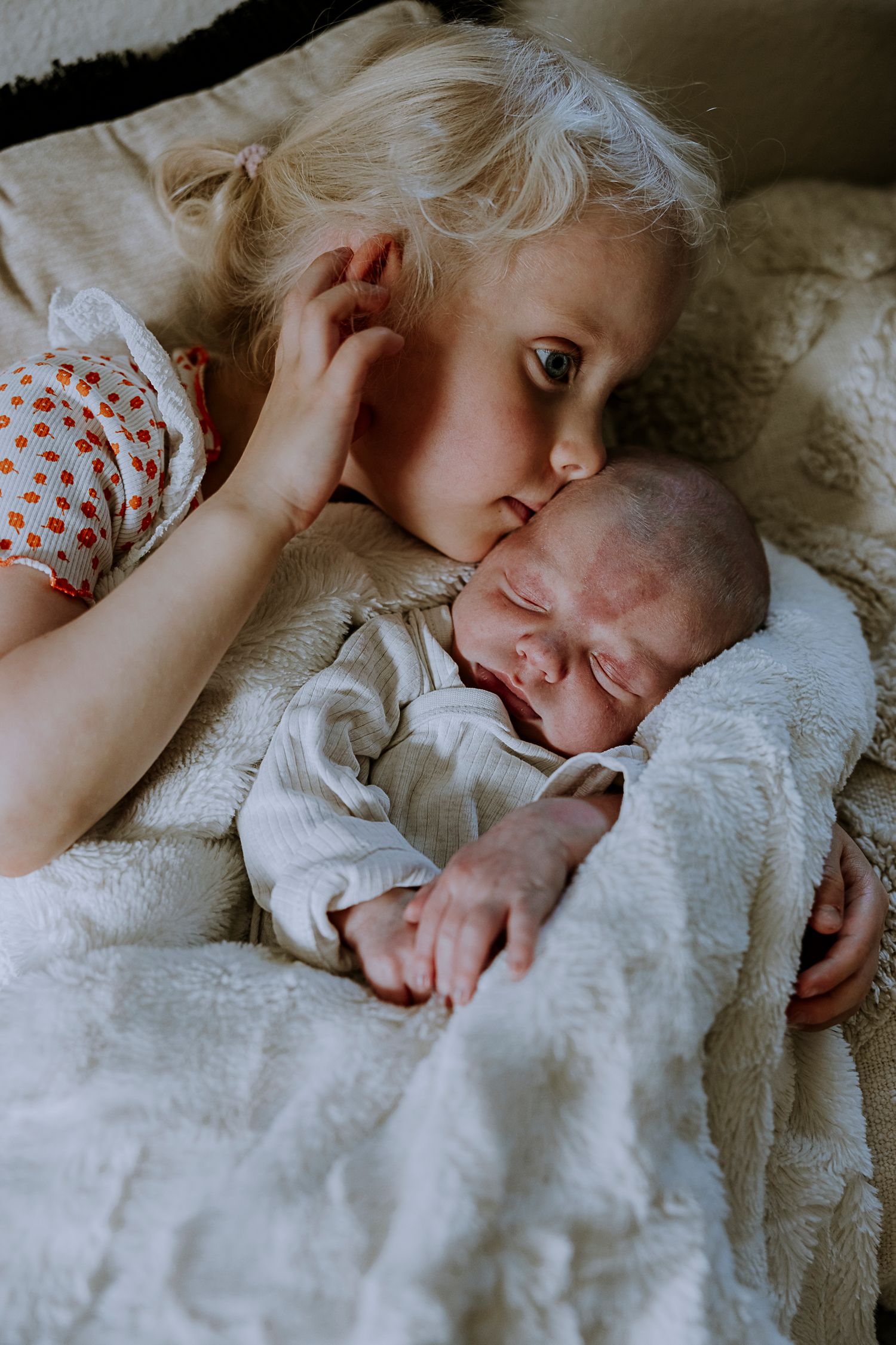 Young child tenderly watching over sleeping newborn sibling on white bedding.