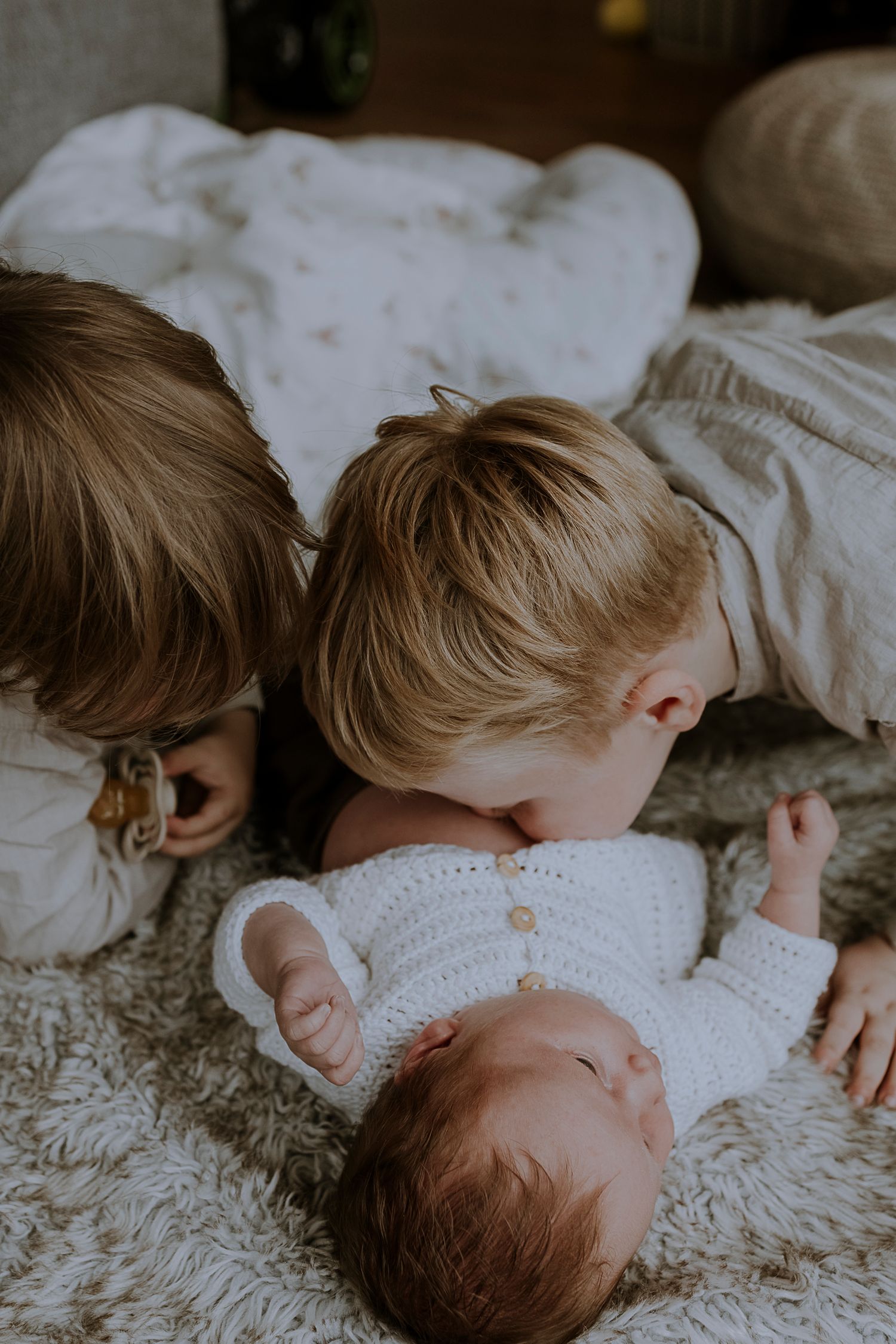 Siblings snuggle close together on white bedding while looking at newborn.