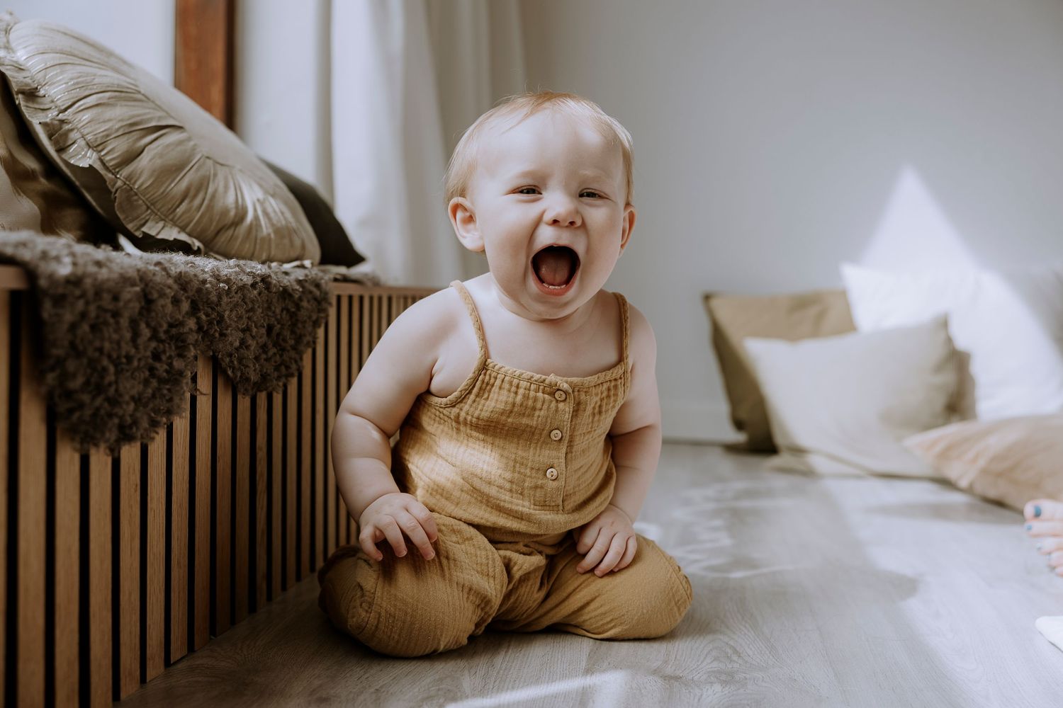 A baby in a mustard romper expressing pure joy while sitting on a bed.
