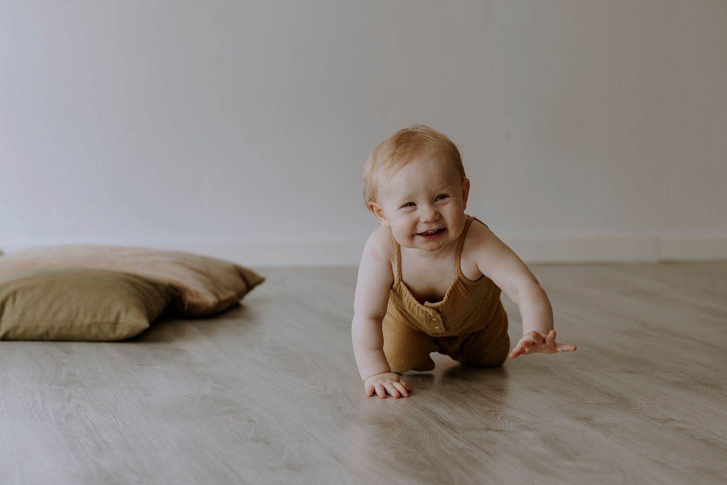 Smiling baby in yellow romper crawls on wooden floor near beige pillow.