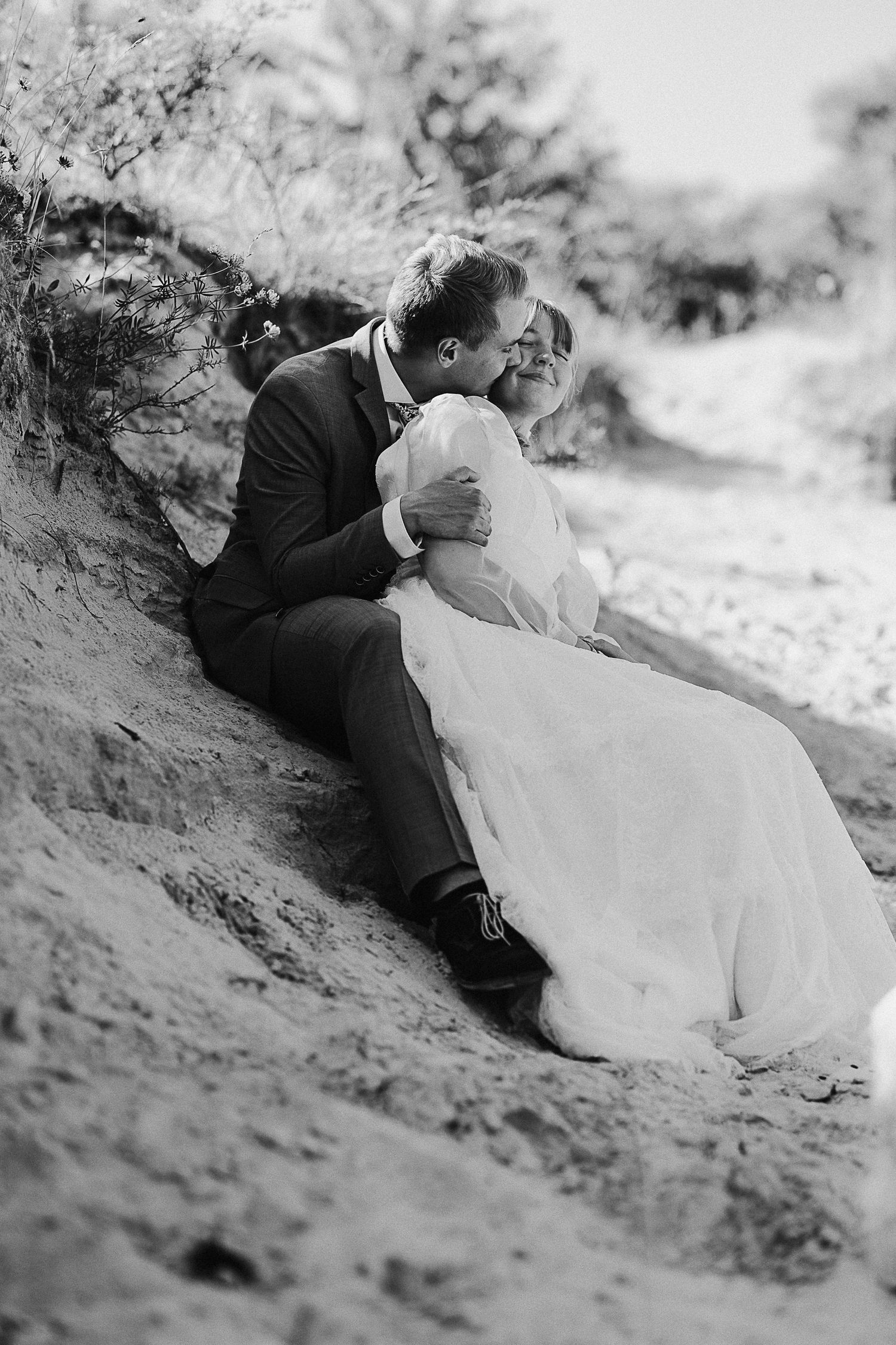 Black and white portrait of a couple in wedding attire sitting together on a hillside.