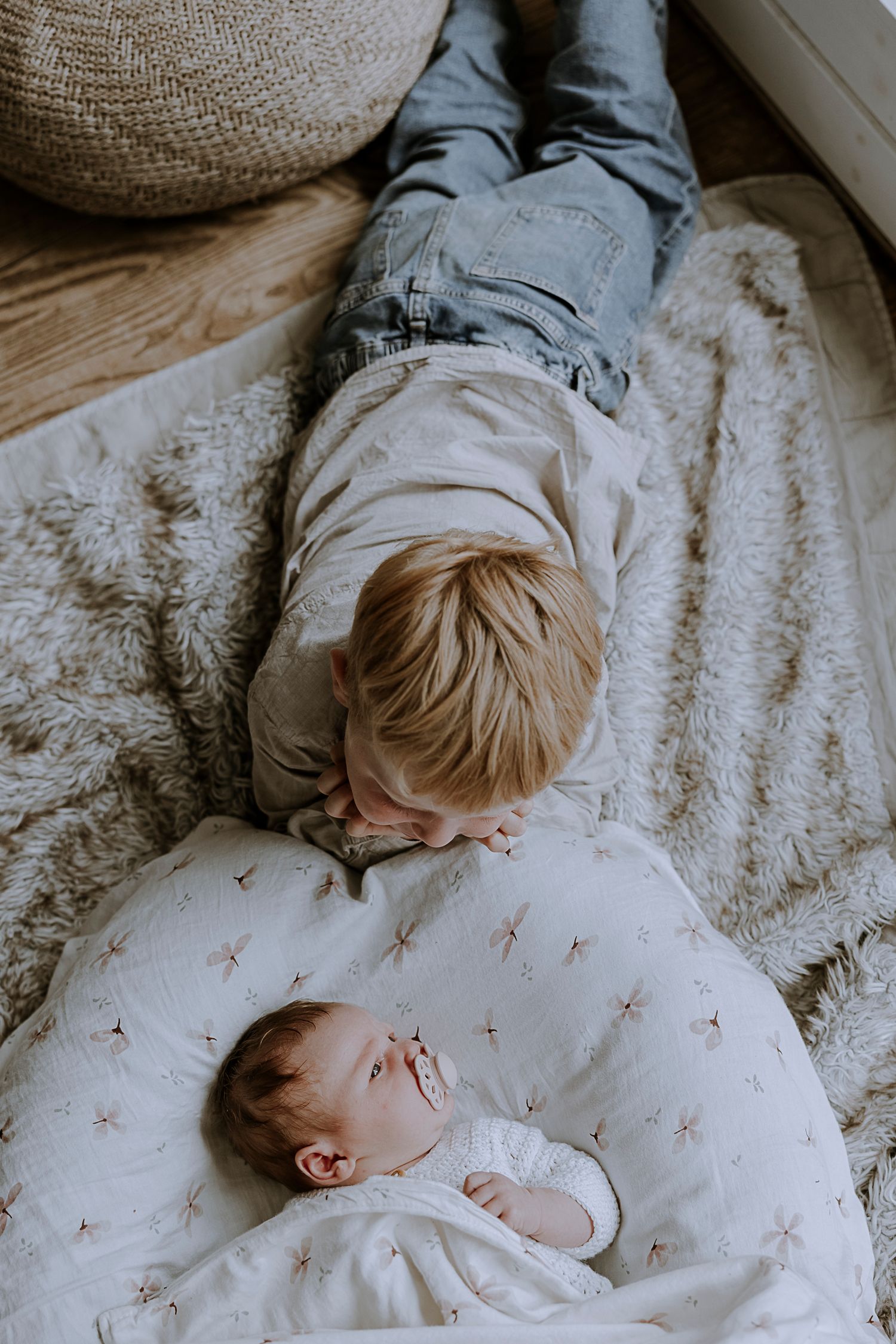 Siblings lying together on a patterned blanket near a woven pouf.