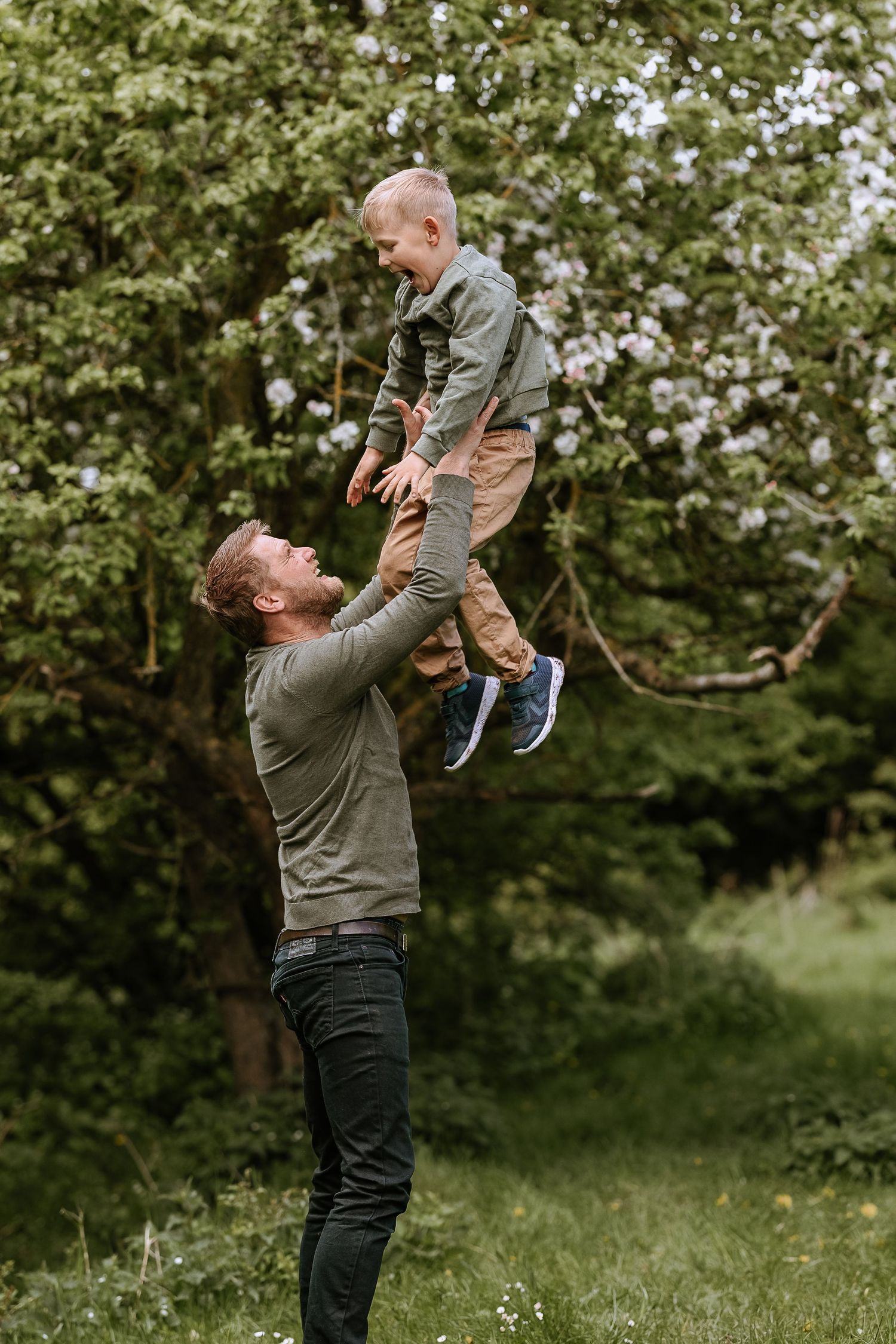 A playful outdoor moment as a parent lifts a small child high into the air against a backdrop of spring trees.