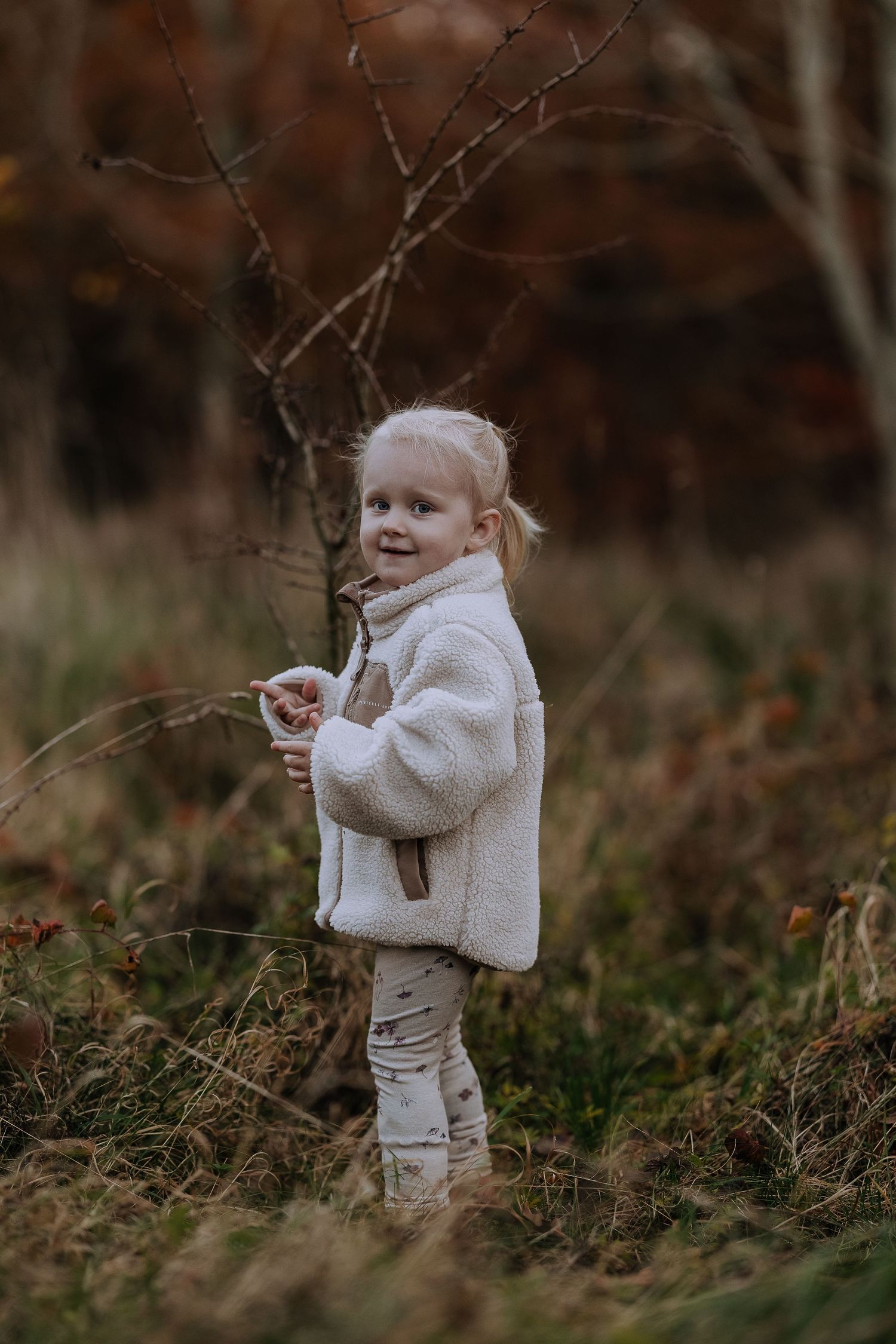 A small child in a fluffy white jacket smiles while playing outdoors among bare branches in autumn.