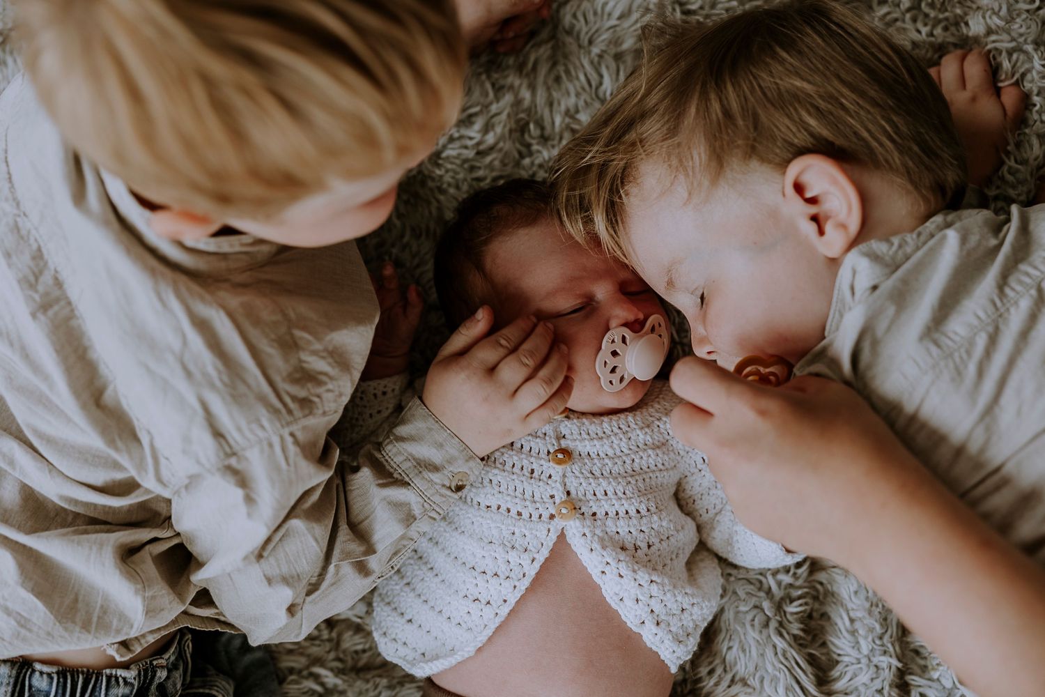 Overhead view of siblings sharing tender moment with baby in white knit sweater.