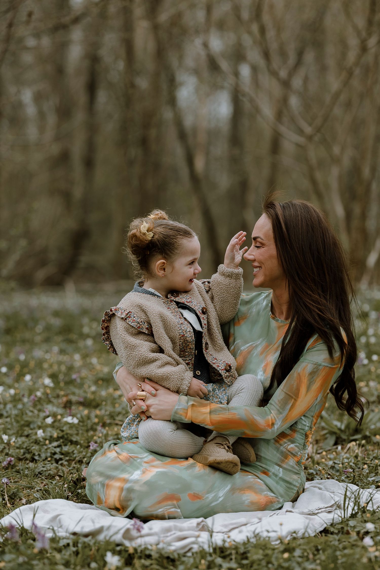 Mother and child share joyful moments on a blanket surrounded by spring wildflowers in a forest setting.