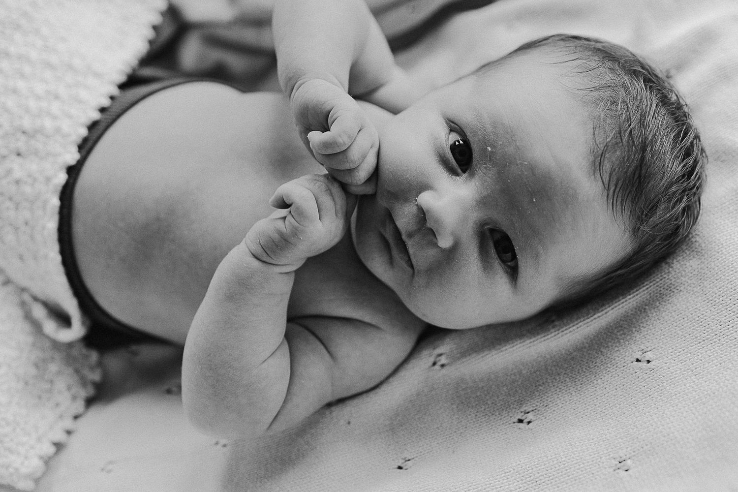 A close-up black and white portrait of a baby with their hand near their face.