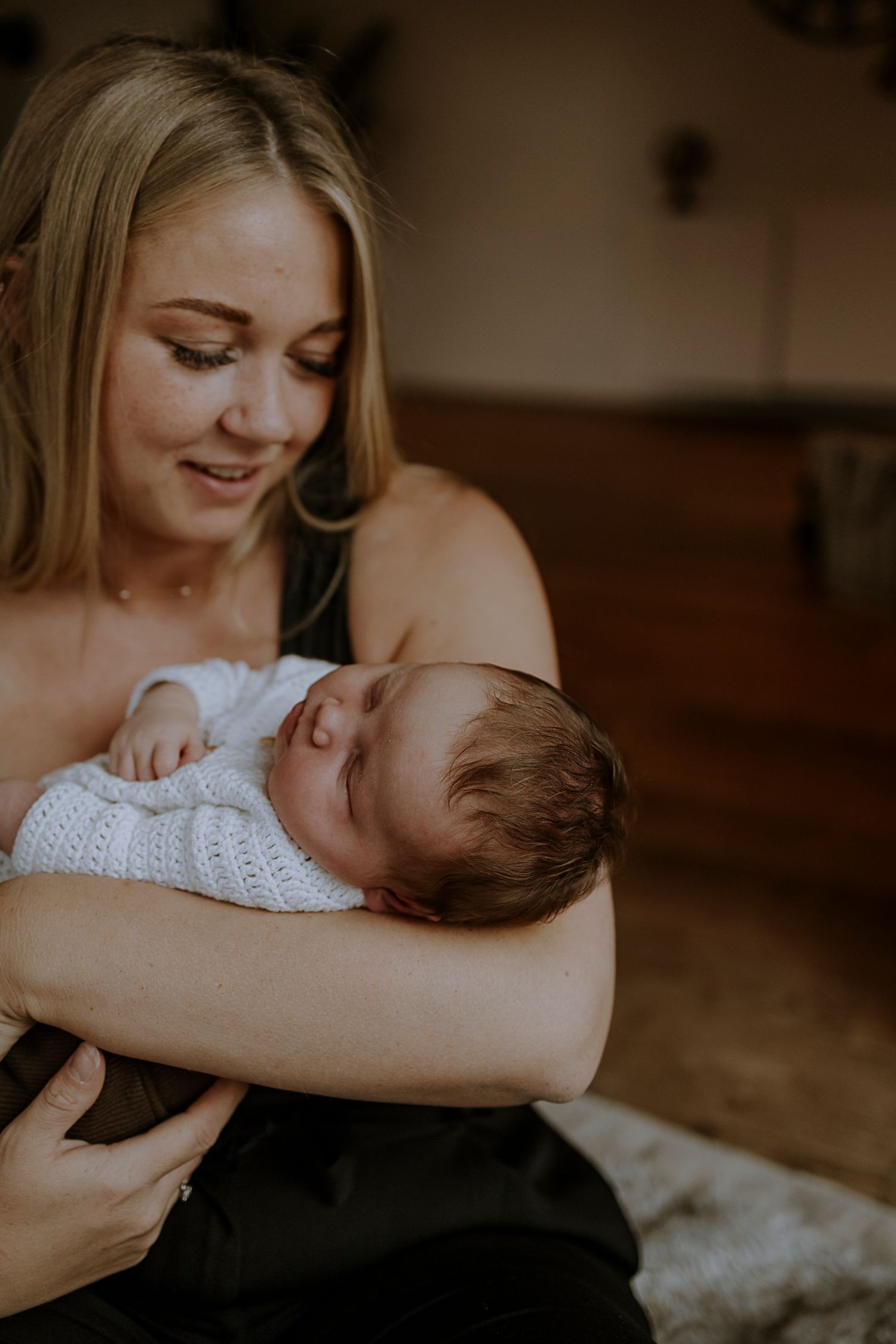 Parent holding sleeping newborn in white knit clothing in warm indoor setting.