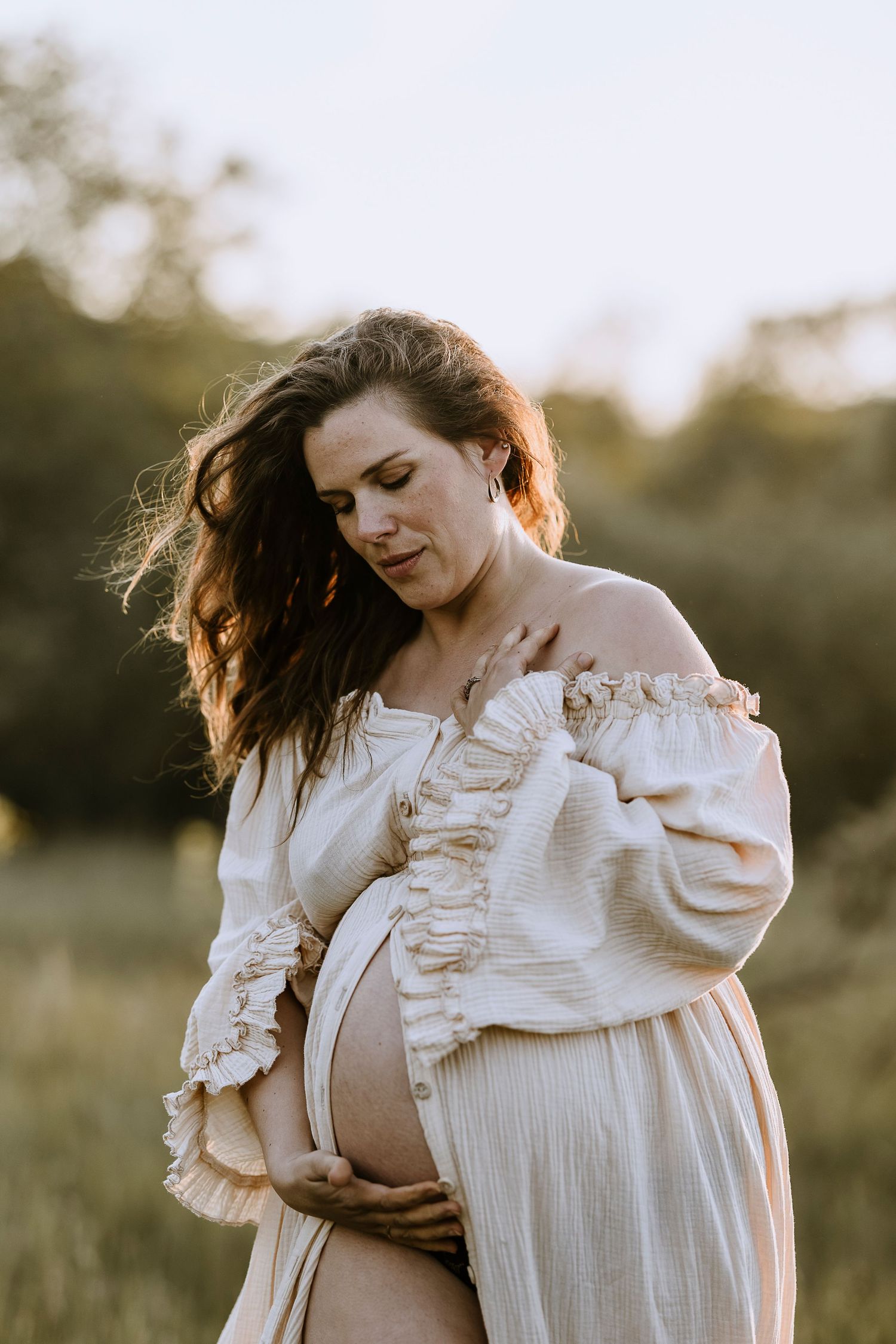 A pregnant figure in a flowing white dress stands in a sunlit field with wind-blown hair at golden hour.