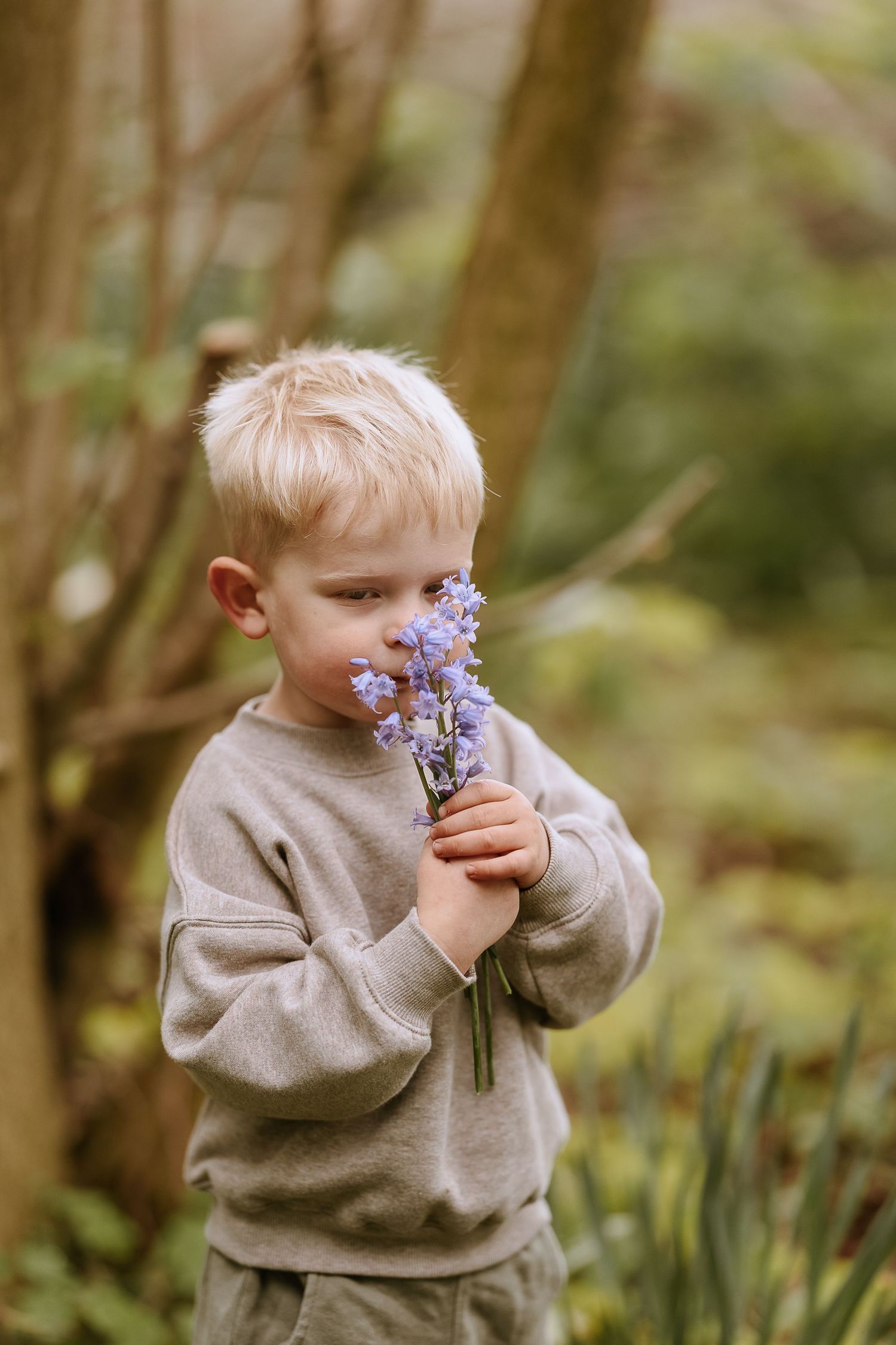 Child in gray sweater smelling purple wildflowers in garden setting.