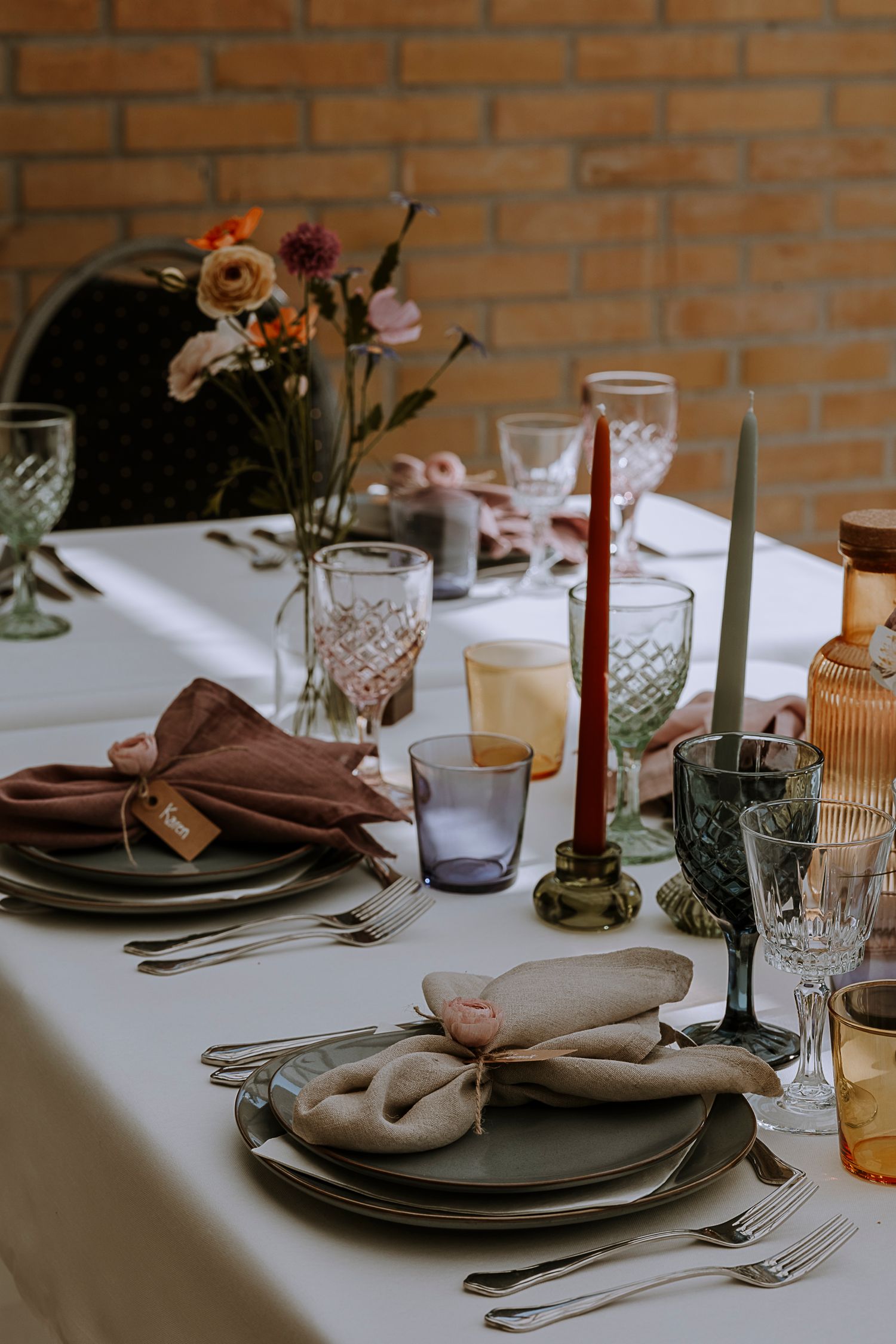 Elegant table setting with crystal glasses, red candles, and floral centerpieces against brick wall.
