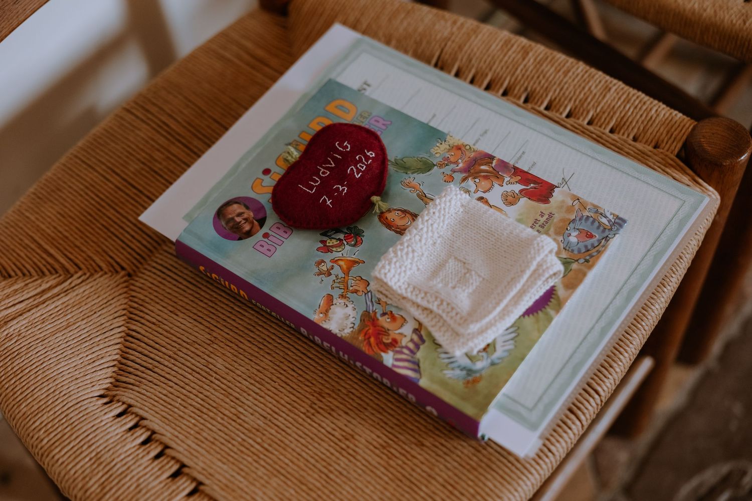 A colorful children's book on a wicker chair with a red heart-shaped ornament and white lace fabric on top.