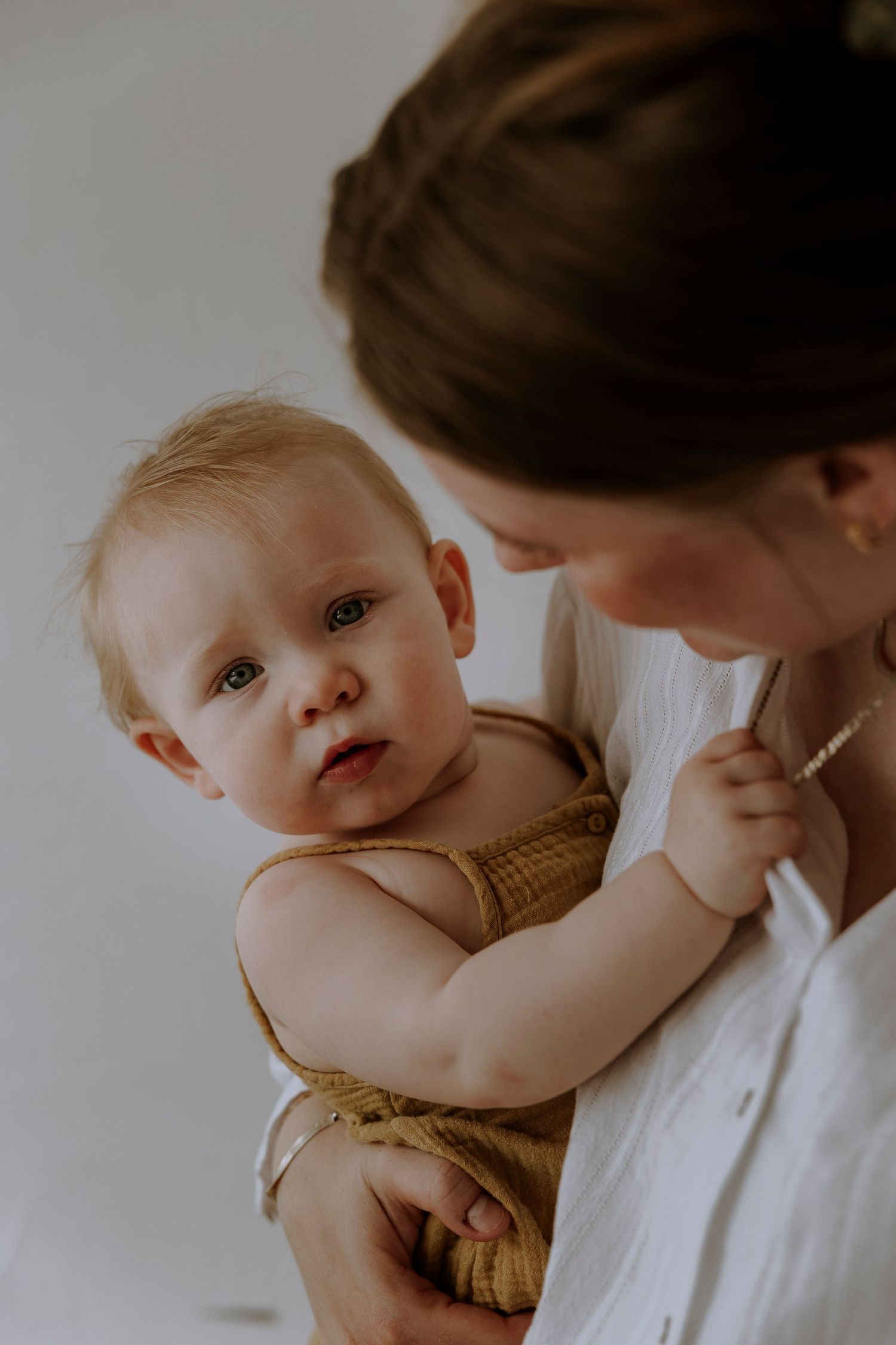 Baby in mustard romper reaches out while being held against white shirt.