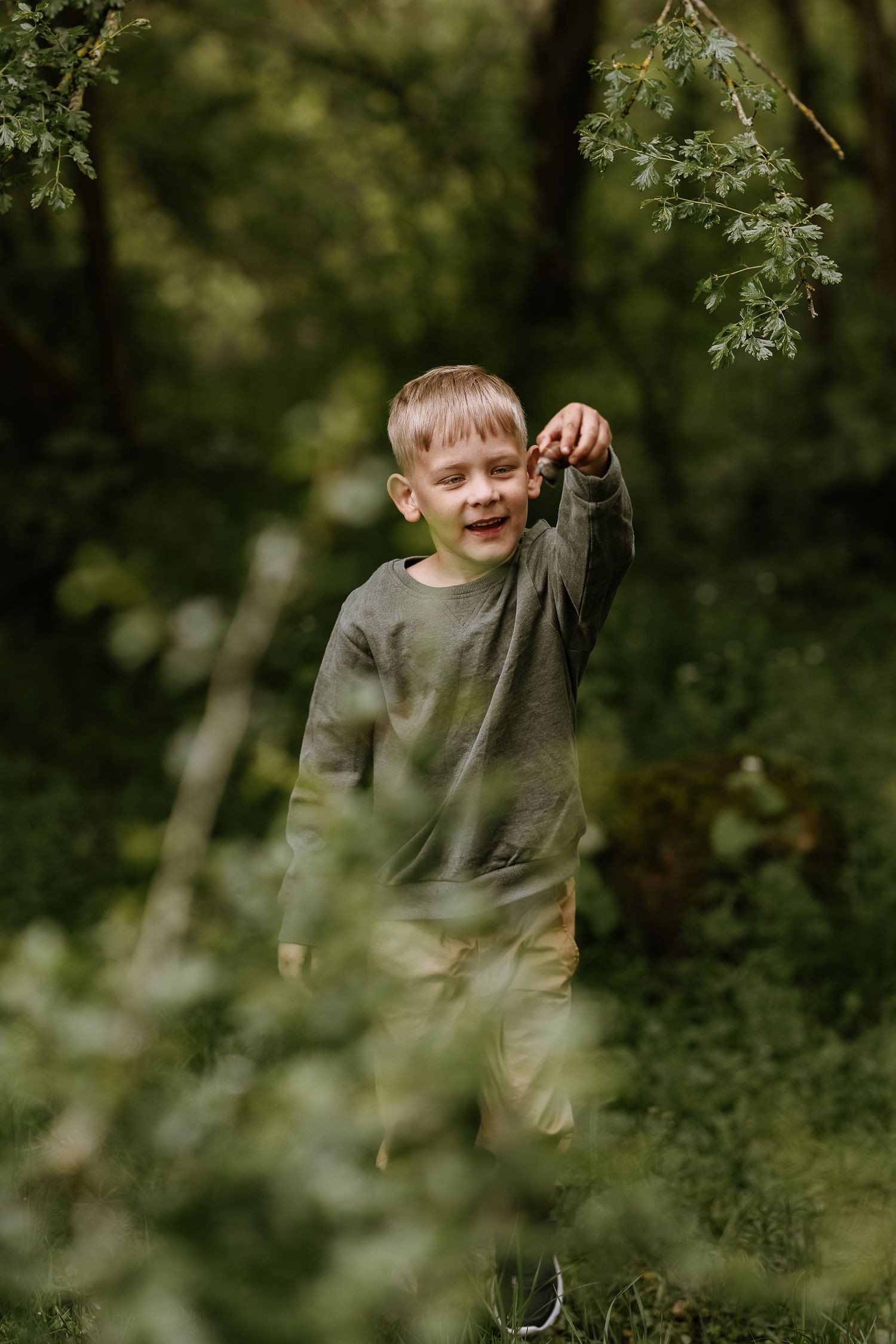 A child in a gray sweater plays among the trees and greenery in a natural outdoor setting.