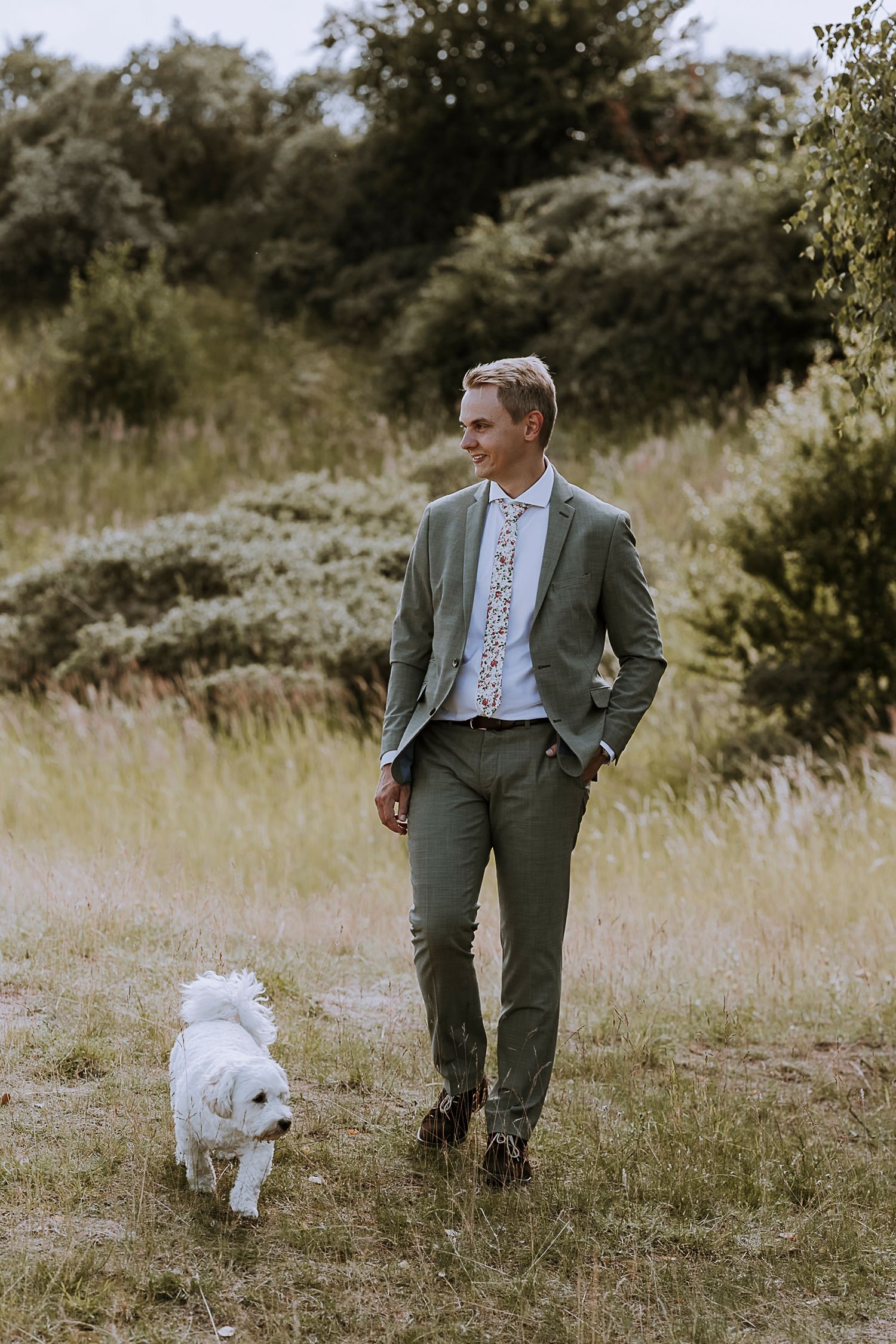 Person in grey suit walking with small white dog through sunny meadow.