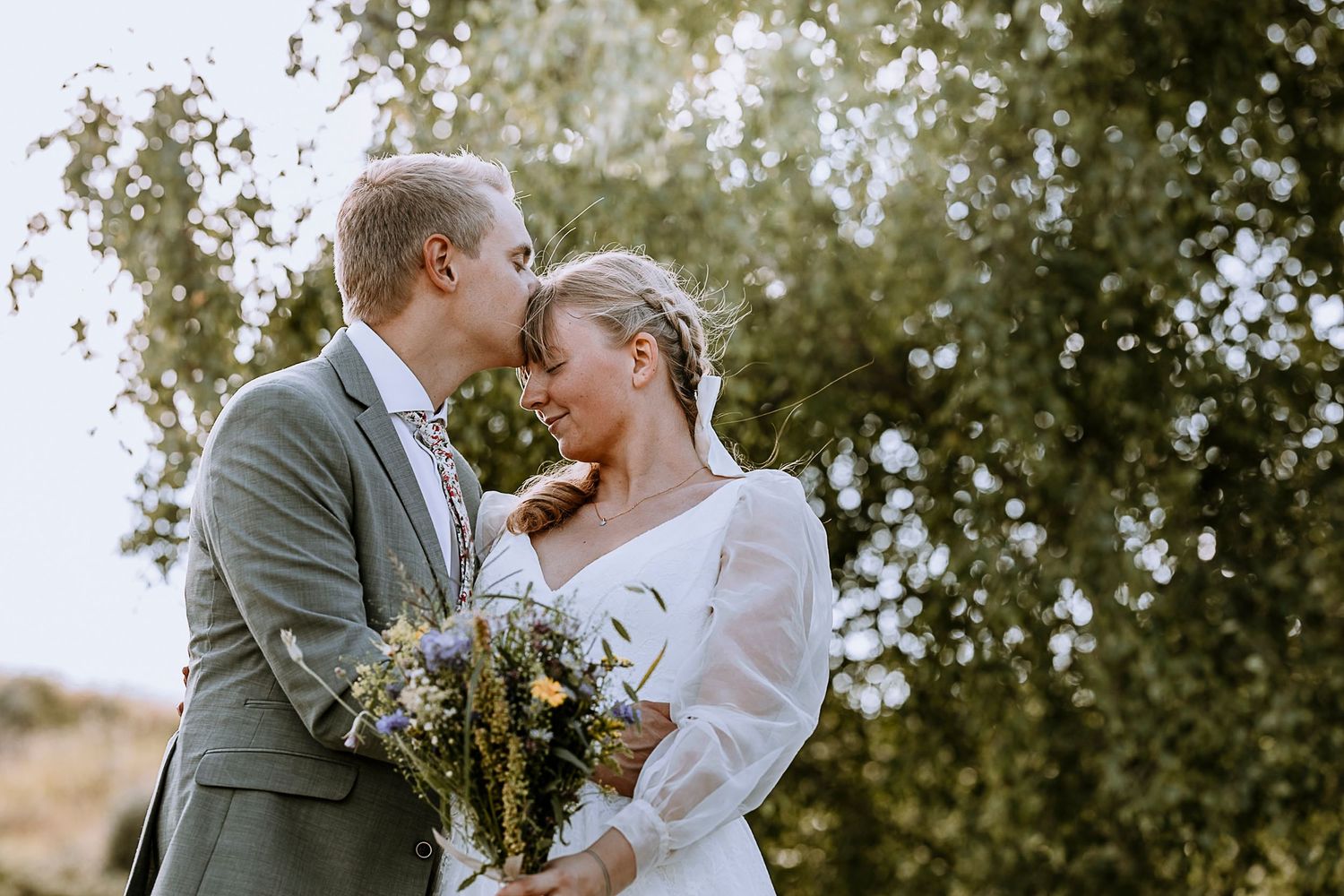 A wedding couple shares an intimate moment outdoors while holding a wildflower bouquet.