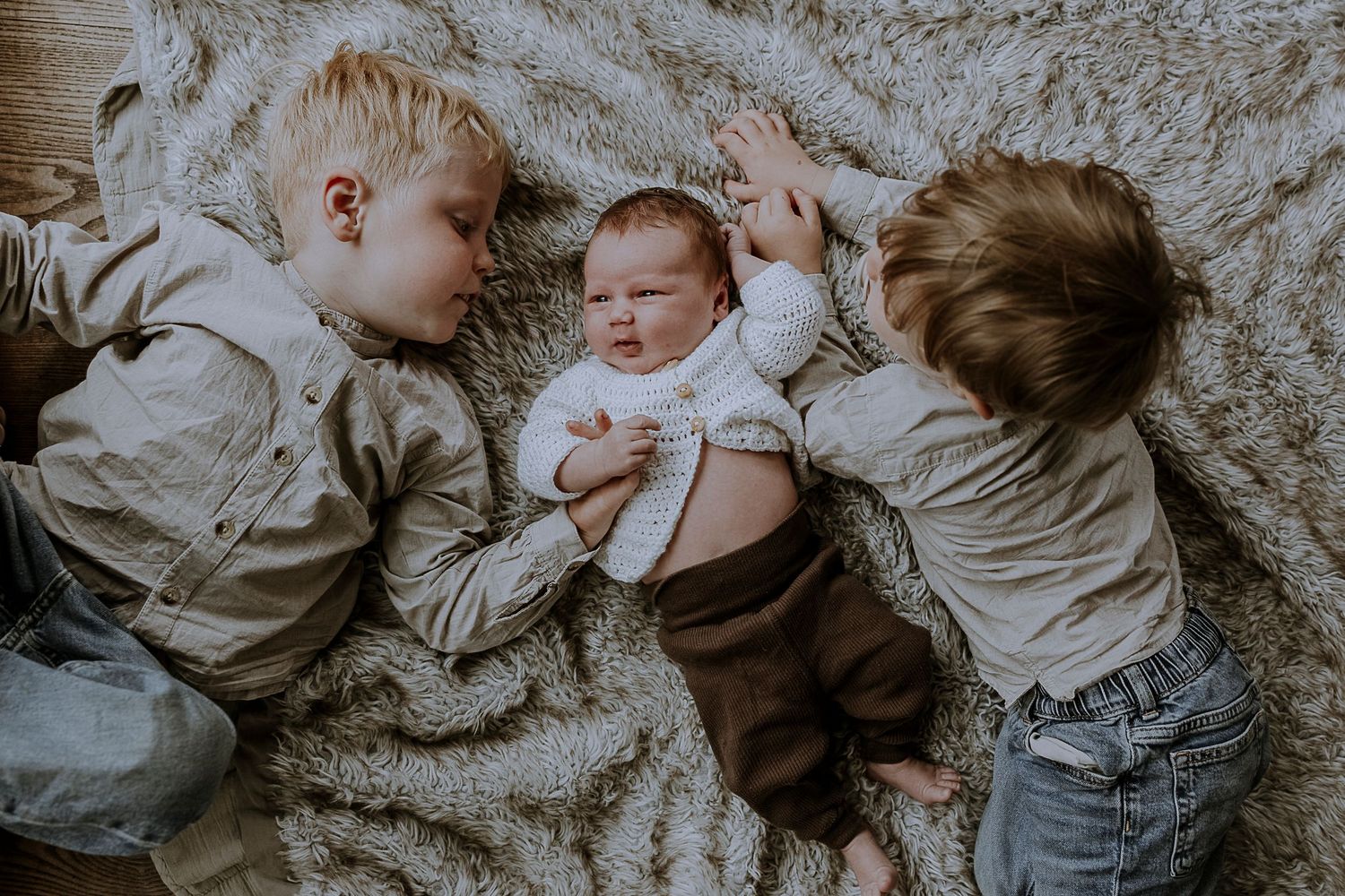 Three children lie together on a plush grey carpet in matching outfits.