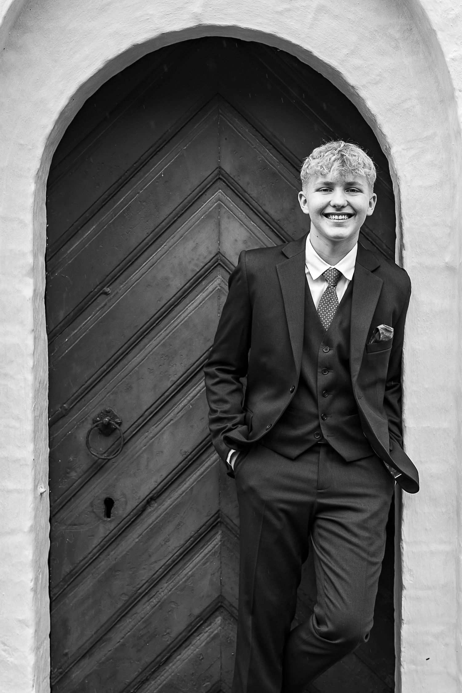 Black and white portrait of a young student in formal suit and tie leaning against an arched wooden doorway.