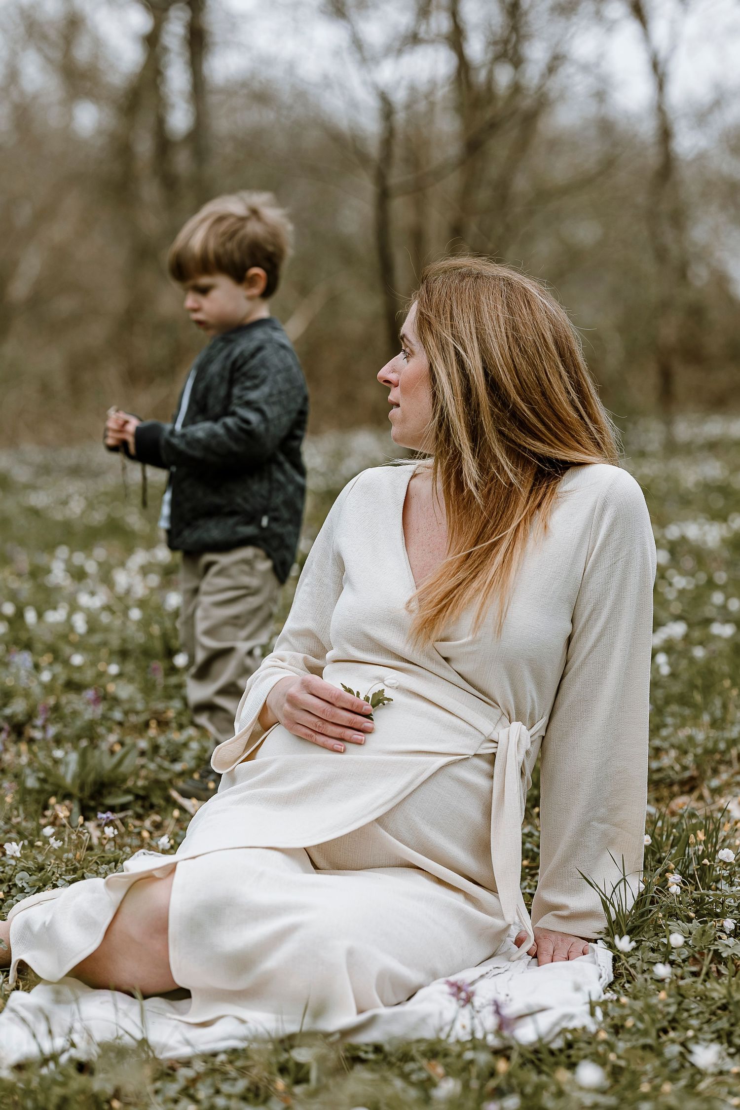 A woman in a white wrap dress sits in a spring meadow filled with small wildflowers while a child stands nearby.