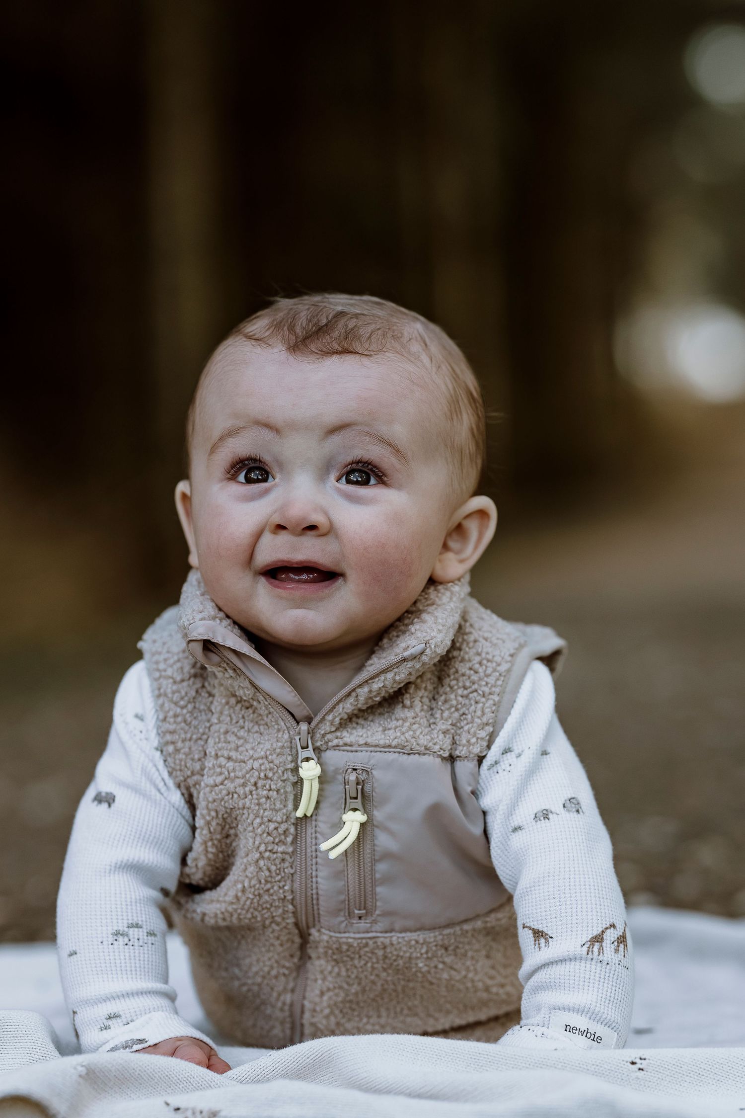 A baby wearing a brown fleece vest and white patterned onesie smiles while laying on a blanket outdoors.