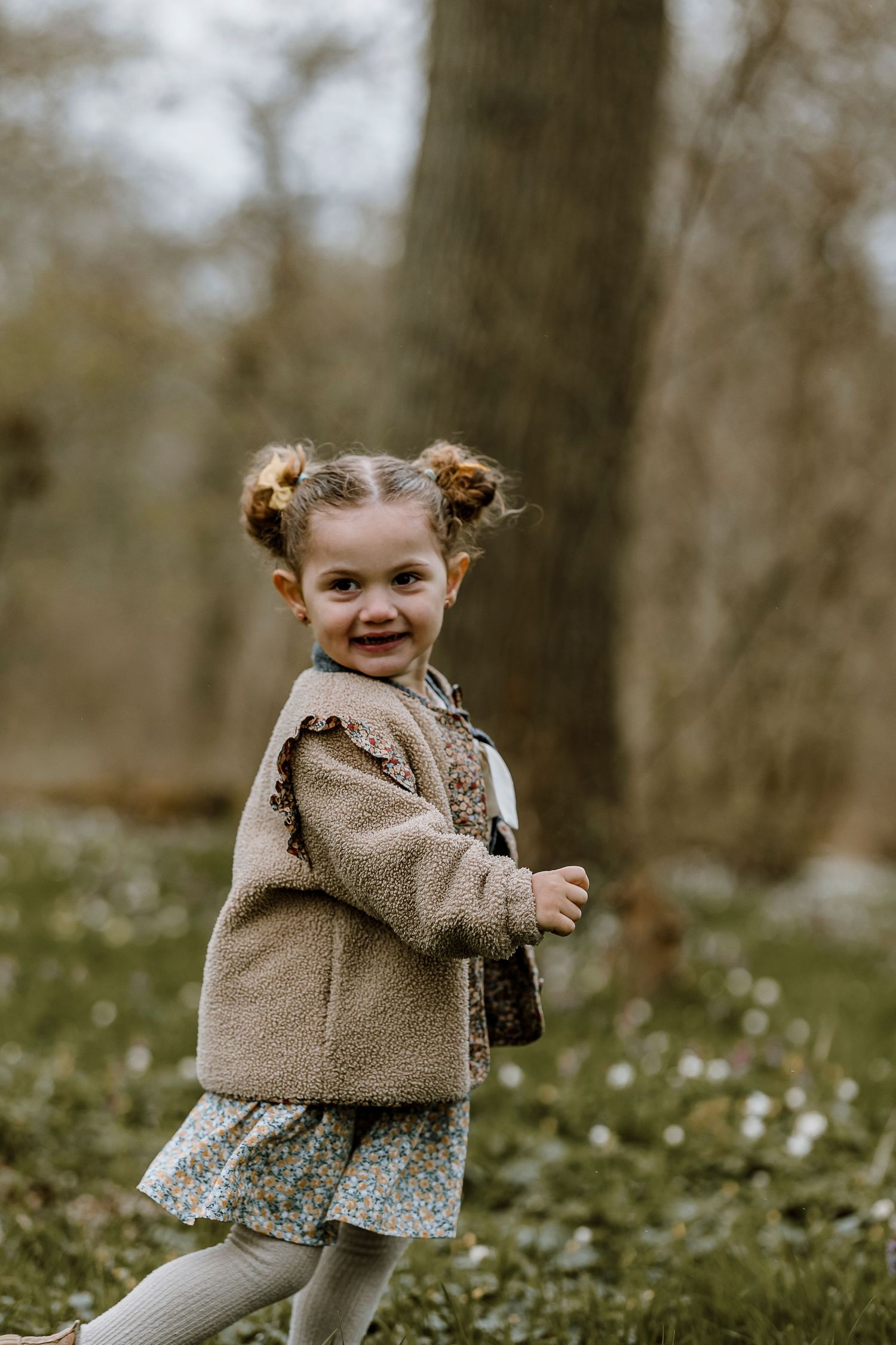A young child in a tan coat and floral dress plays outdoors in a wooded setting on an autumn day.