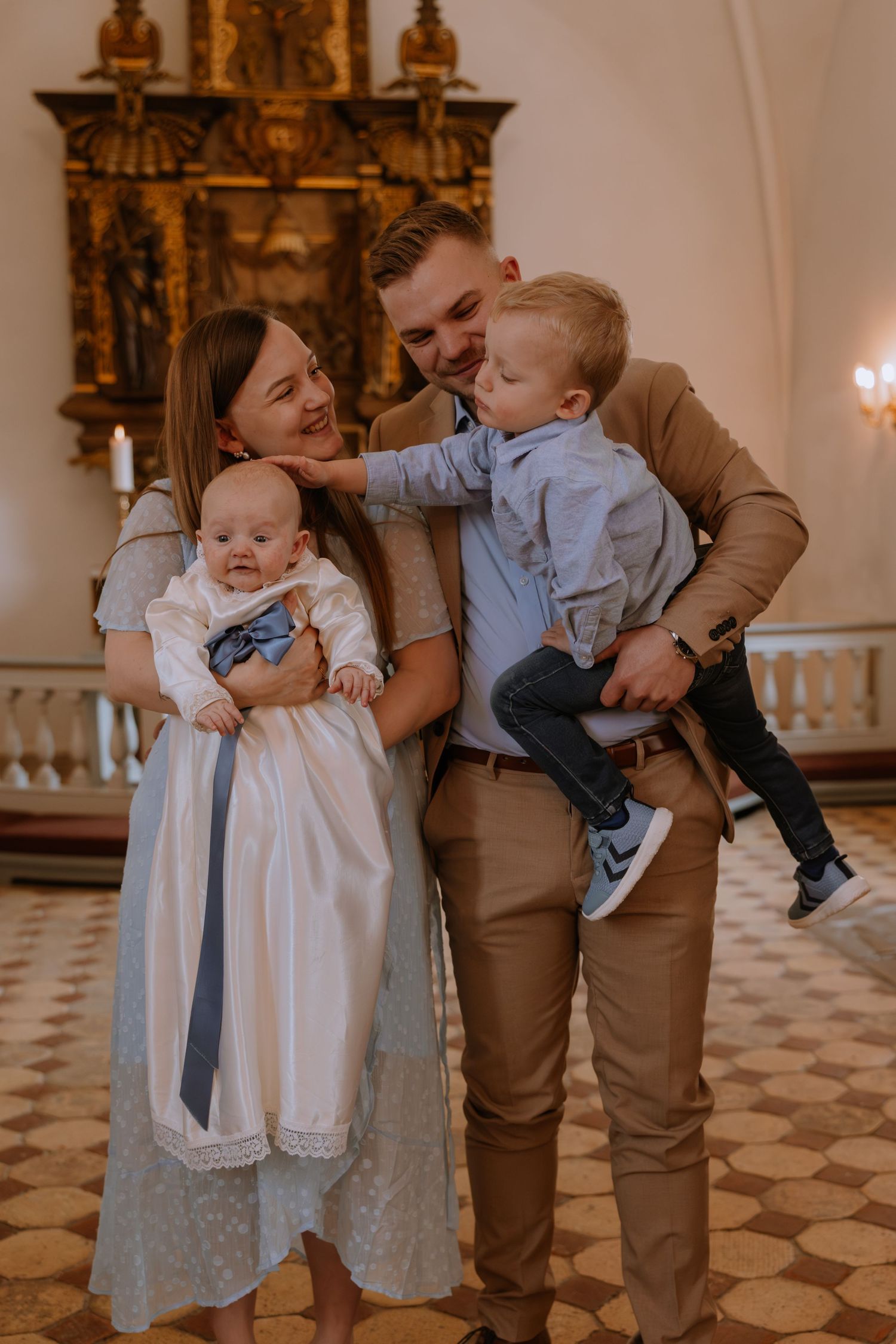 Family of four posing in a church during a baptism ceremony, parents holding two young children dressed in formal attire.