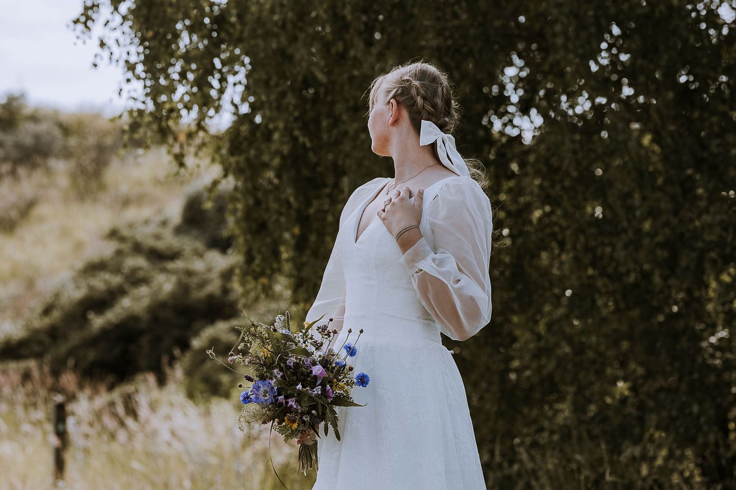 A bride in a white dress holds a wildflower bouquet against a natural outdoor backdrop.