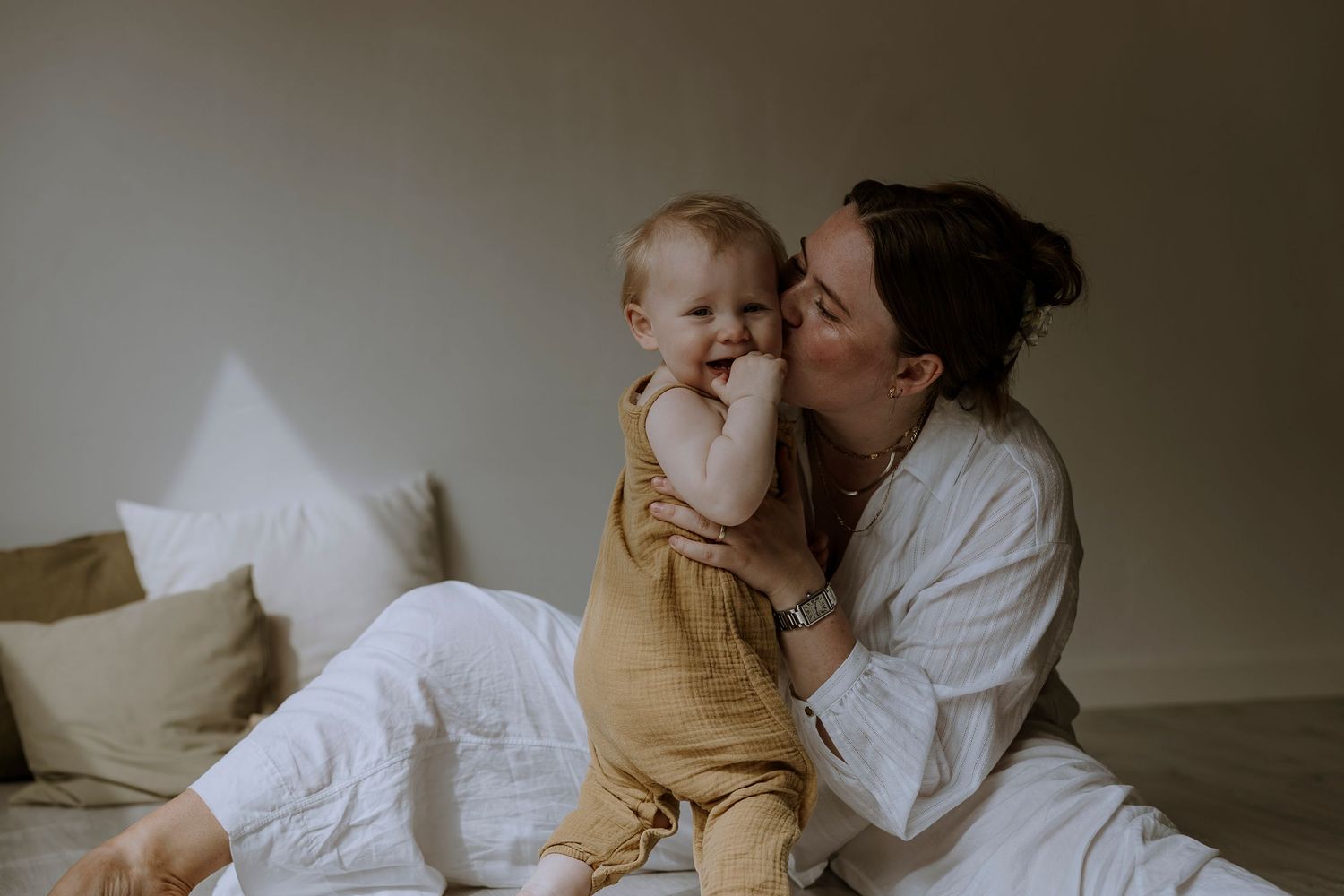 Baby in yellow romper shares tender moment with parent on white bedding.