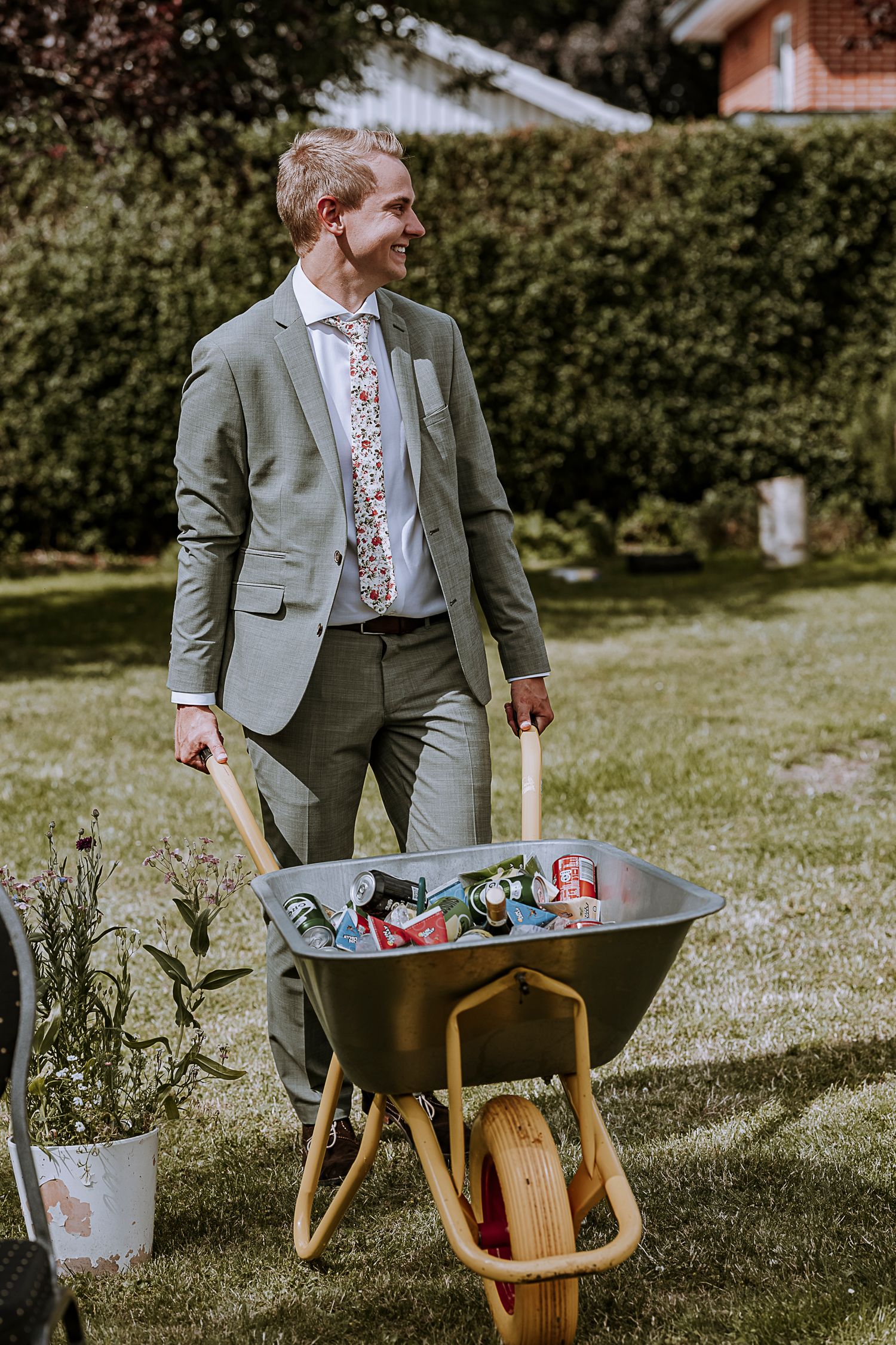 Suited guest carrying wheelbarrow of drinks across sunny lawn at outdoor wedding.
