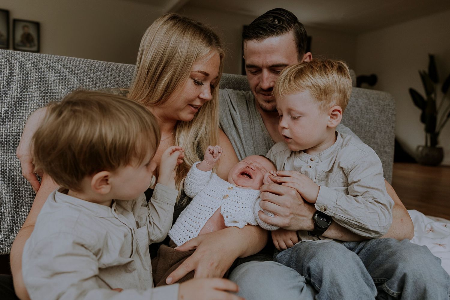 Family shares intimate moment on grey couch while bonding with newborn baby.